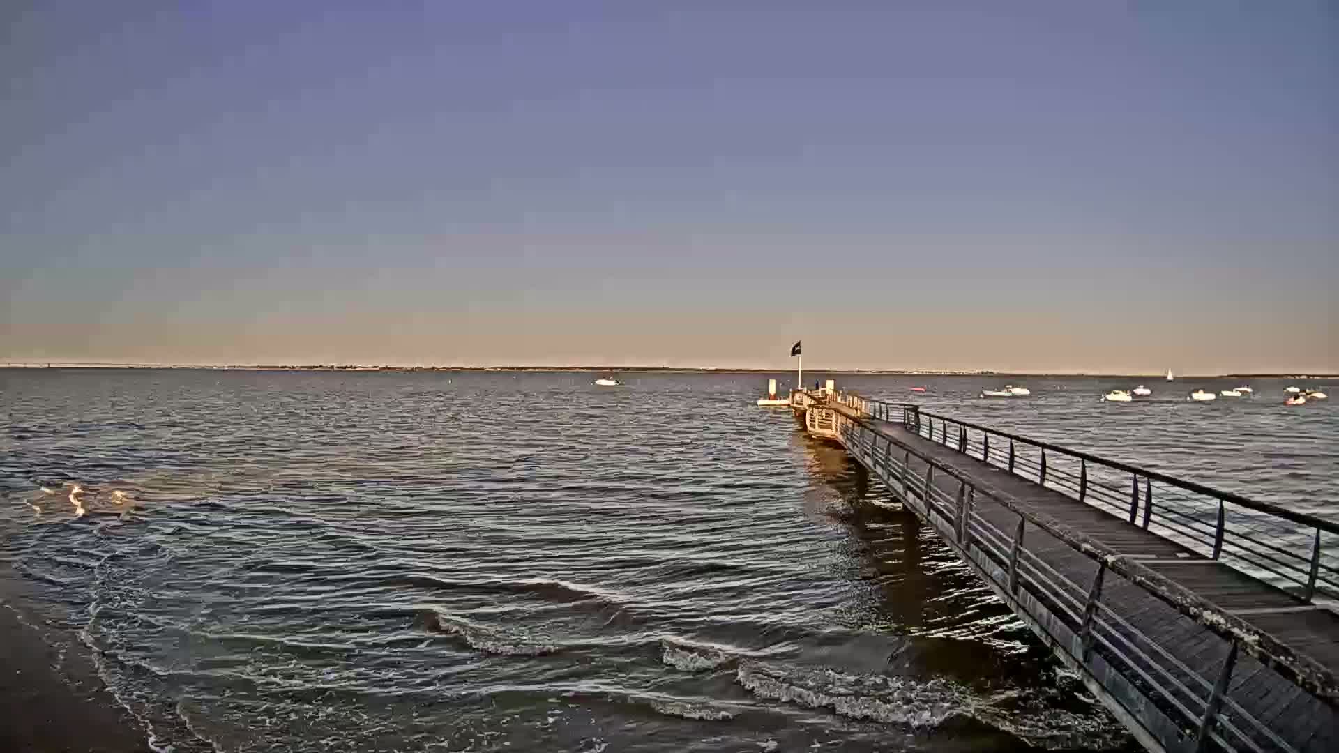 A long pier extends into calm, slightly wavy water under a clear, pale-blue sky, with several small boats visible in the distance.