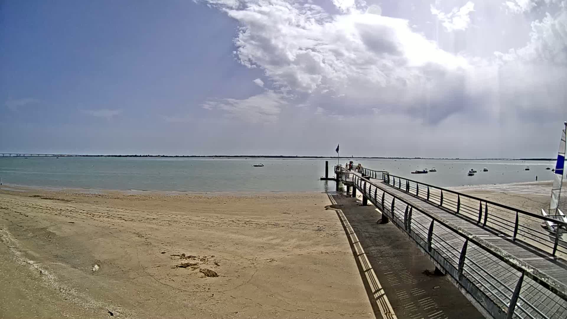 A sandy beach leads to a long pier extending over calm, light-green water under a partly cloudy sky.