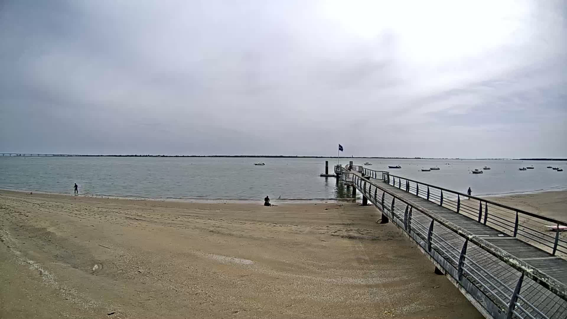 A long pier extends from a sandy beach into calm, grey water where several small boats are anchored under a cloudy sky.