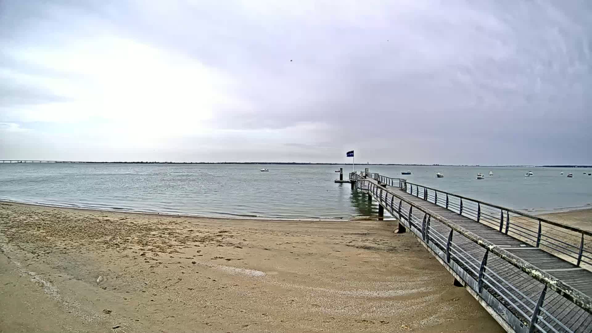 A long wooden pier extends from a sandy beach into calm, gray water under an overcast sky, with a few boats visible in the distance.
