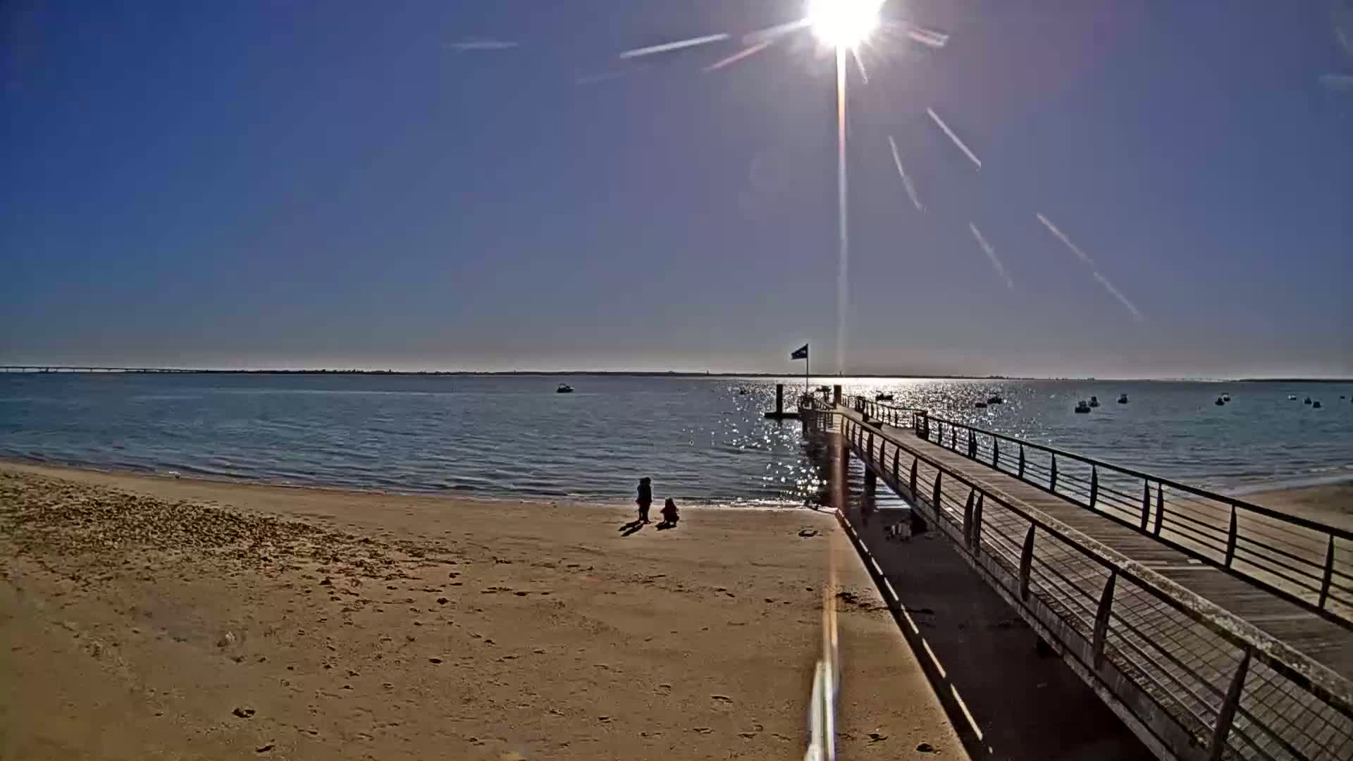 A sandy beach stretches to a long pier extending into a calm, sunny bay with several boats visible in the distance.