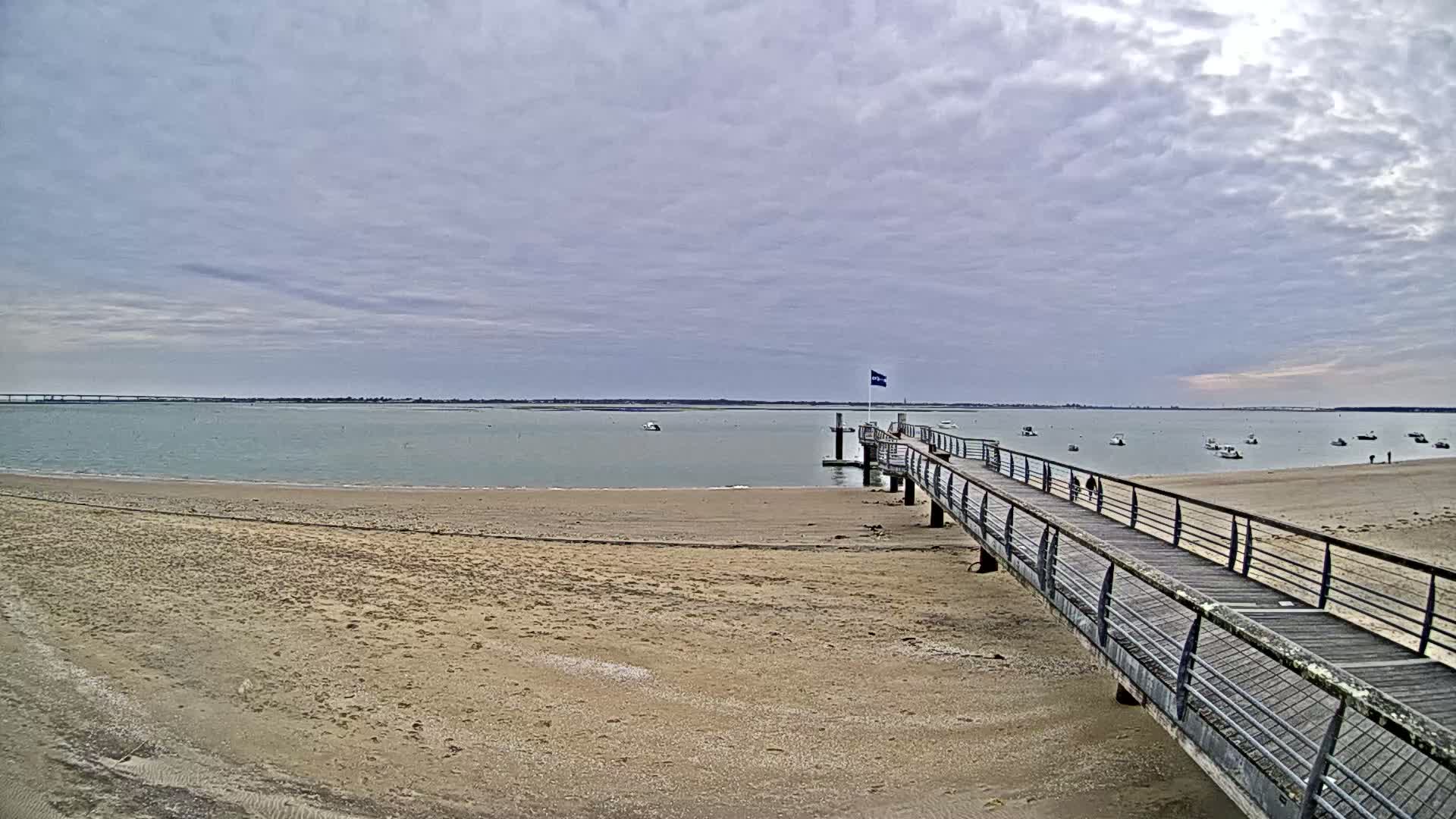 A long pier extends from a sandy beach into calm, light-green water under a cloudy sky, with several small boats visible in the distance.