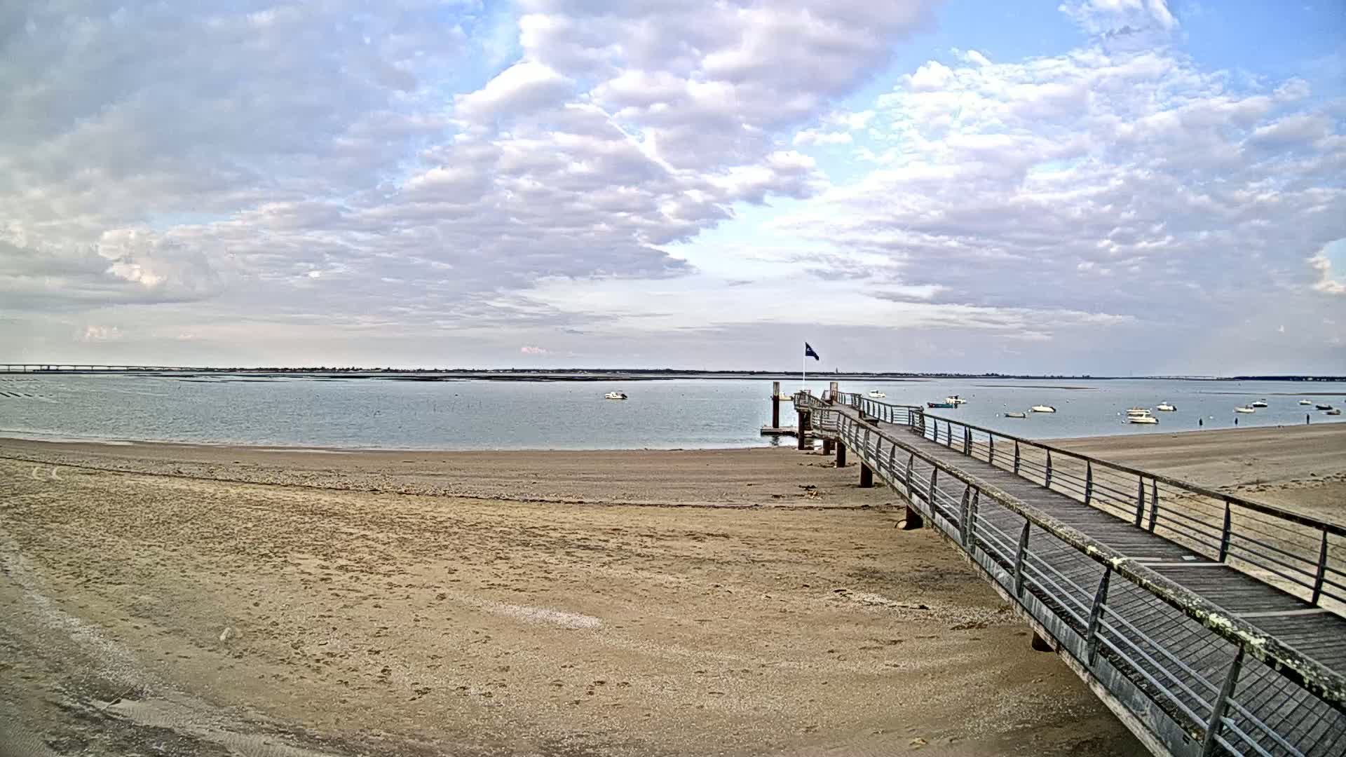 A long wooden pier extends from a sandy beach into a calm body of water under a partly cloudy sky, with several small boats visible in the distance.
