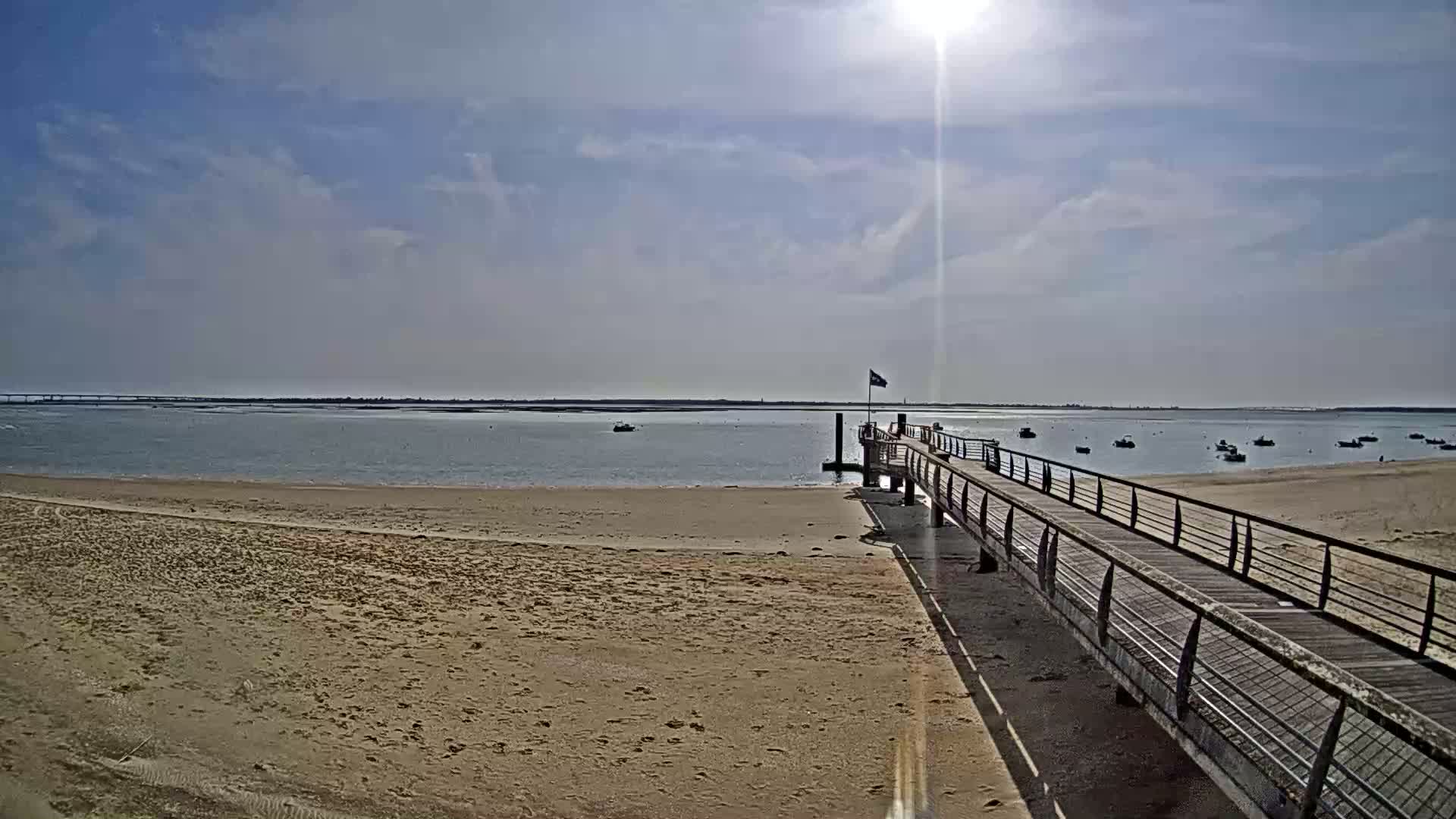 A long pier extends from a sandy beach into calm, shallow water under a bright, sunny sky with a few scattered clouds.