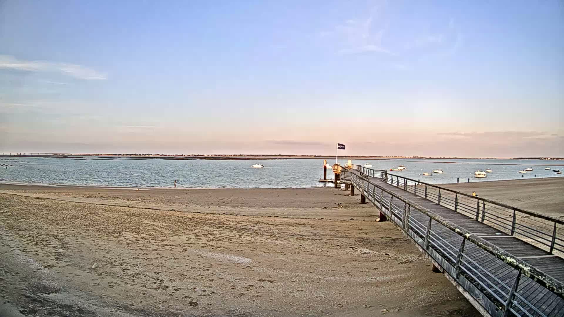 A long wooden pier extends from a sandy beach into calm water, with several small boats visible in the distance under a clear, pale sky.