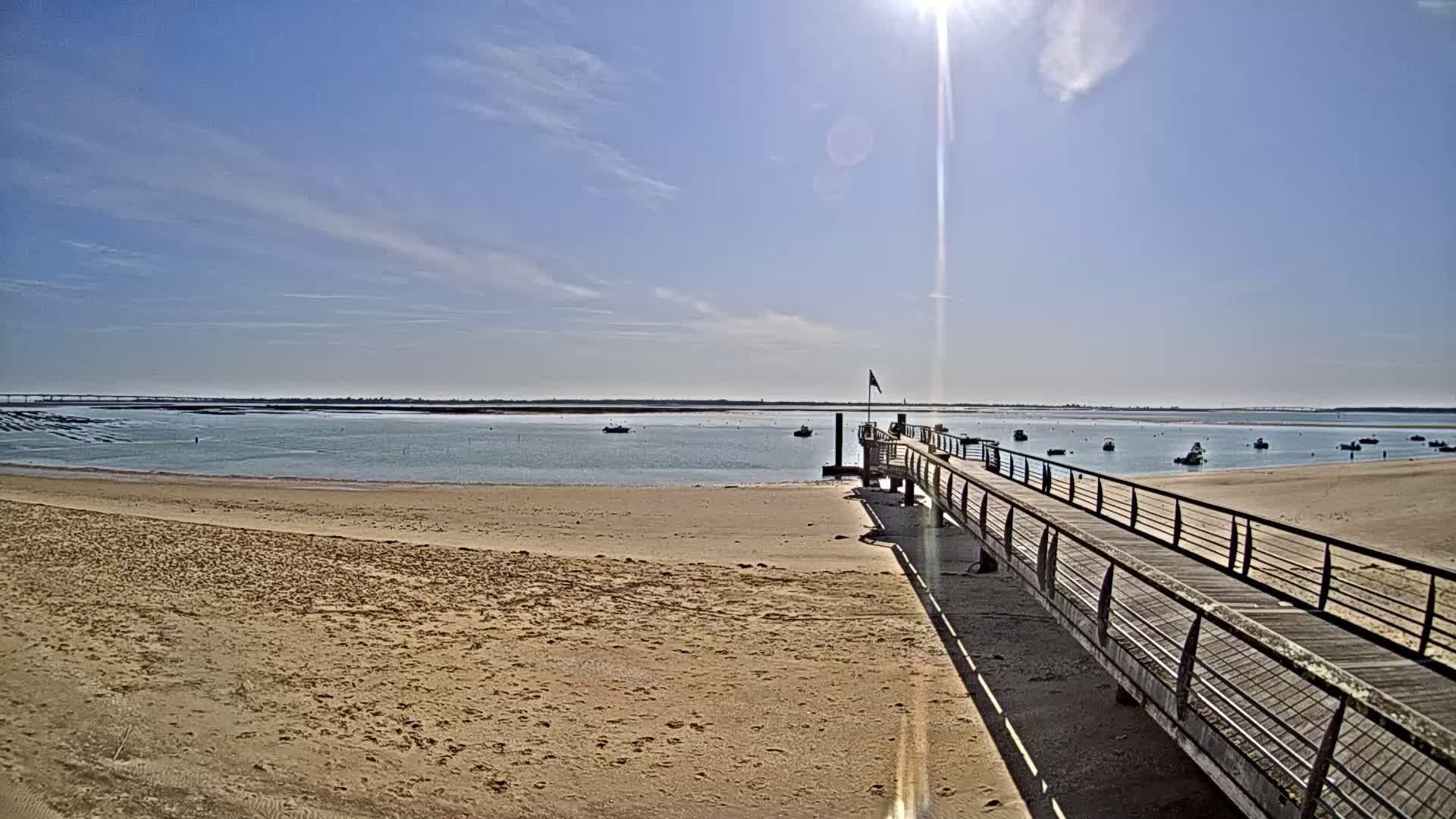 A long pier extends from a sandy beach into a calm, sunny bay with several small boats at anchor.