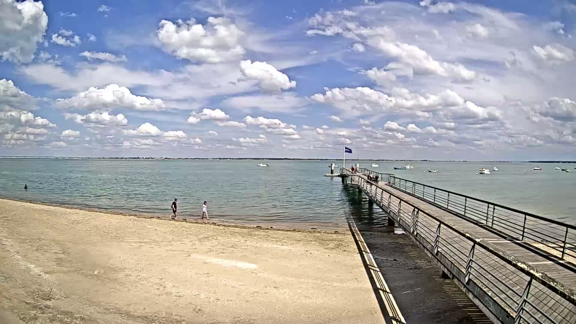 A sandy beach with a few people on it leads to a long pier extending into a calm body of water under a partly cloudy sky.