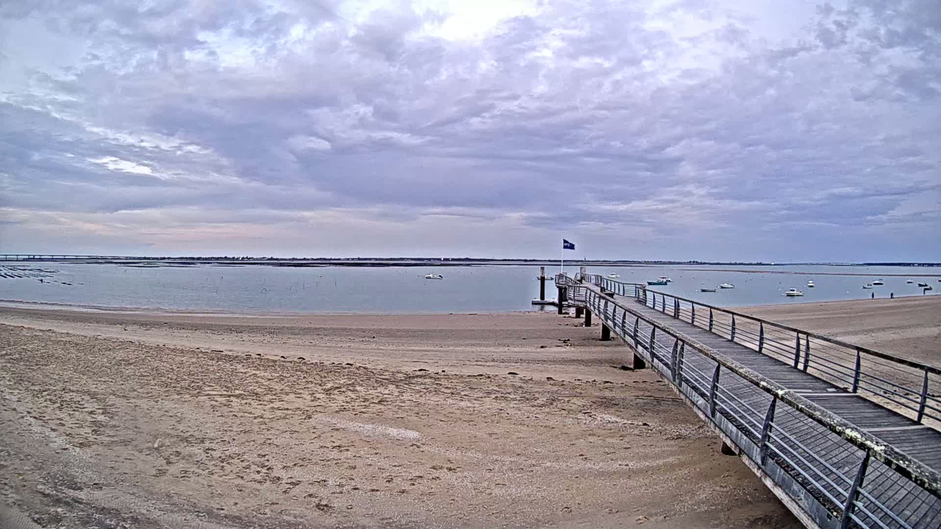 A long wooden pier extends from a sandy beach into calm, shallow water under a cloudy sky, with several small boats visible in the distance.