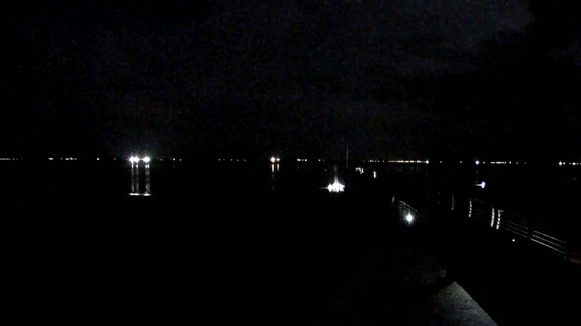 A dark night scene shows a body of water with distant lights on the opposite shore, and a dimly lit pier or dock in the foreground.