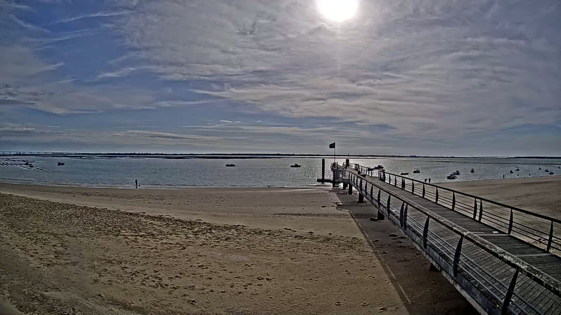 A long wooden pier extends from a sandy beach toward a calm body of water with several small boats under a mostly sunny sky.