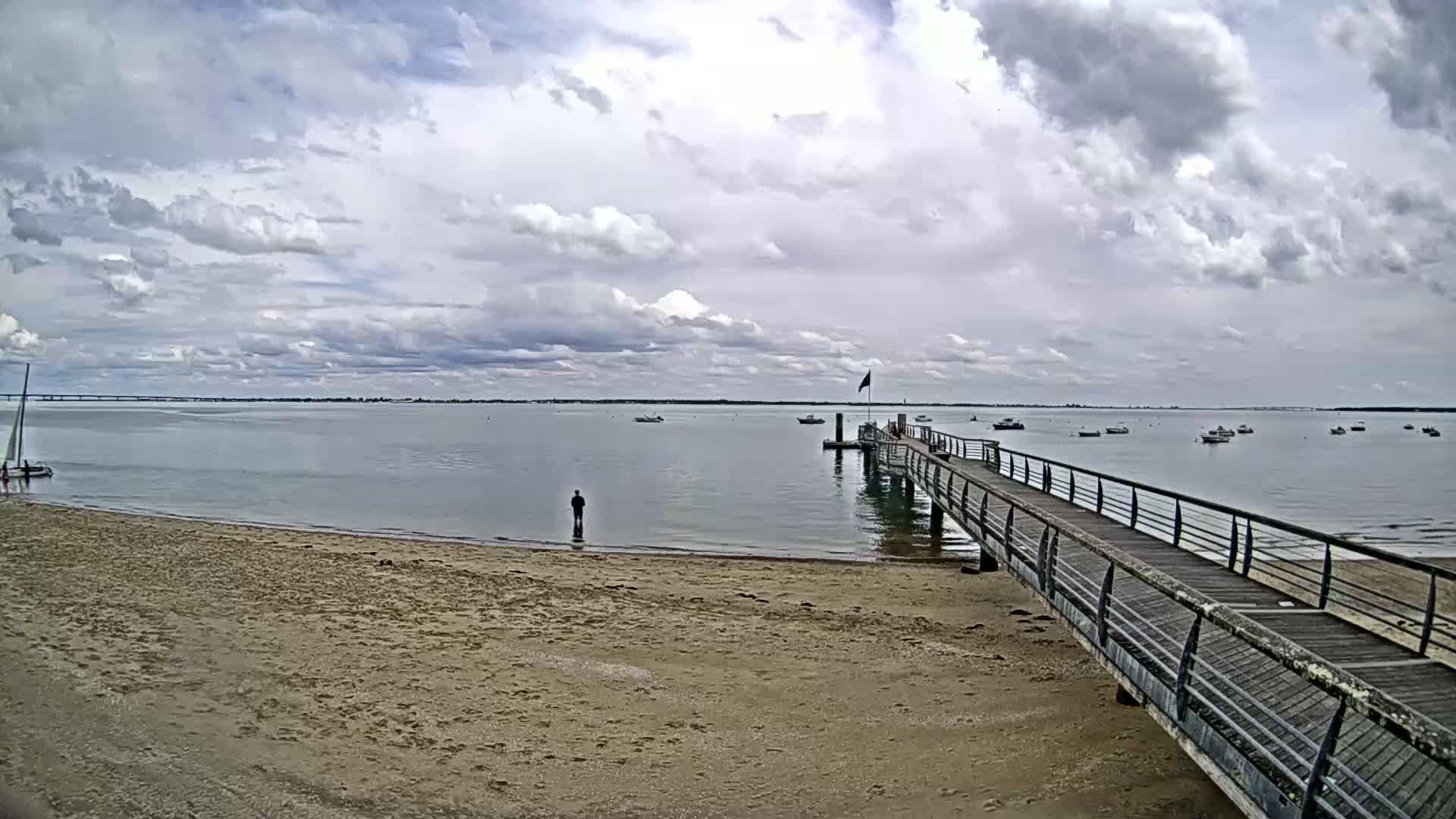 A sandy beach with a wooden pier extends into a calm body of water under a cloudy sky, several small boats are visible in the distance.