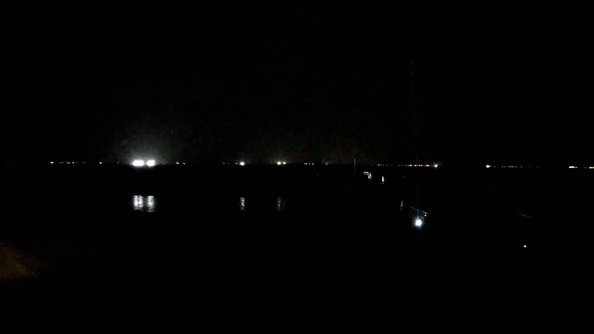 A dark, nighttime scene shows a body of water reflecting faint lights from a distant shoreline.