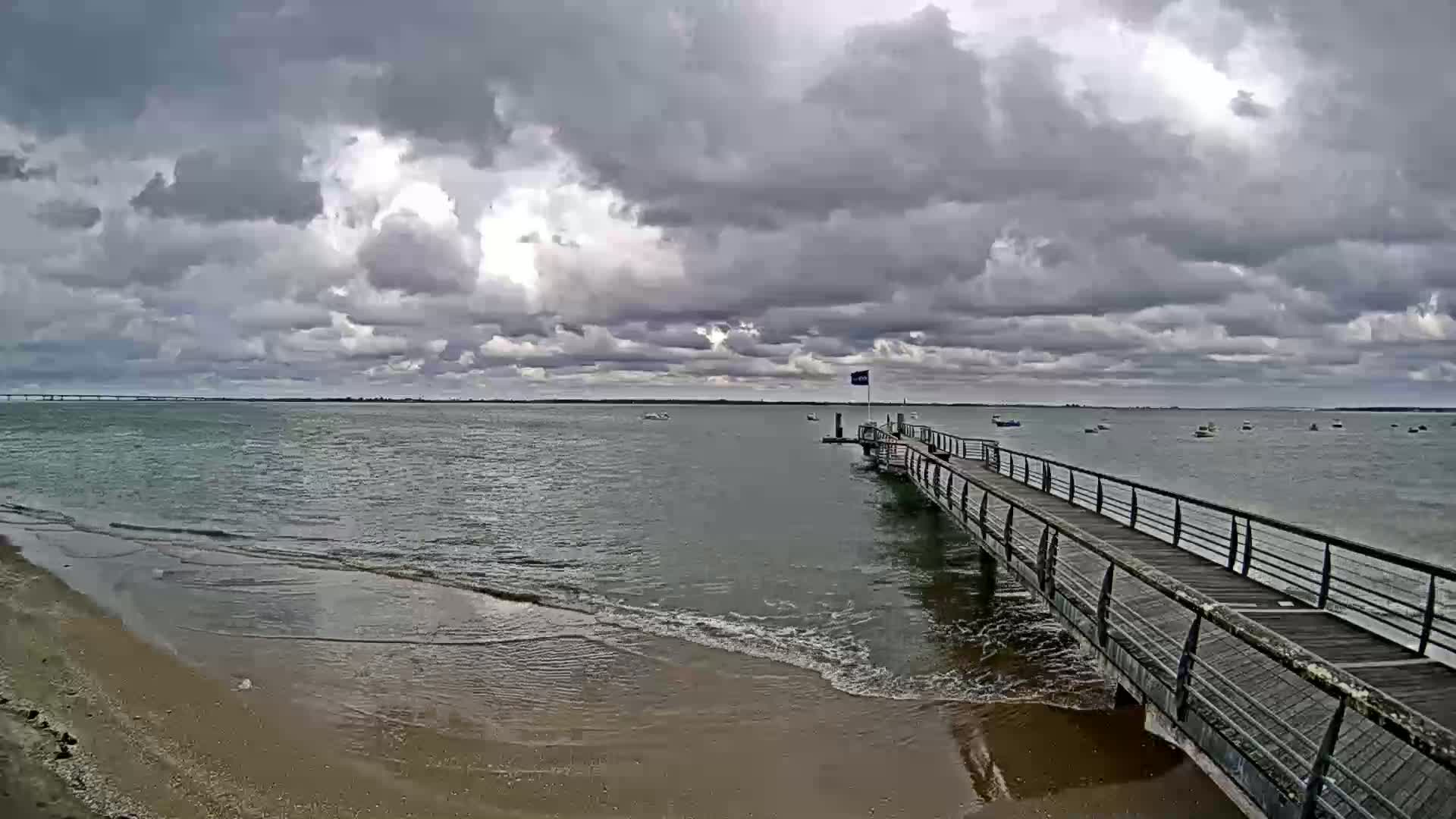 A long pier extends over calm, gray water under a cloudy sky, with a sandy beach visible in the foreground.