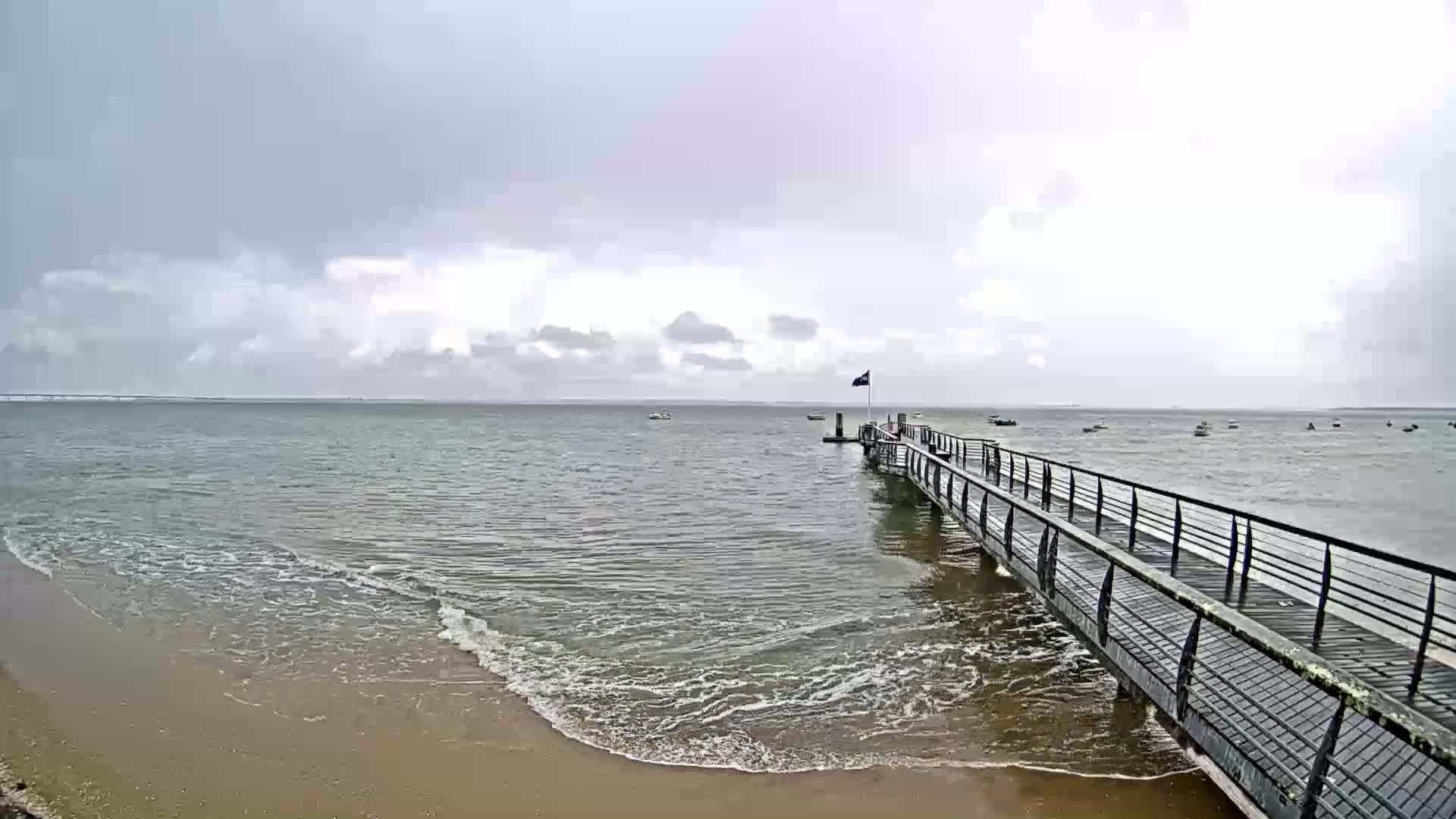 A long pier extends into a calm, gray ocean under an overcast sky, with small waves lapping at a sandy shore.