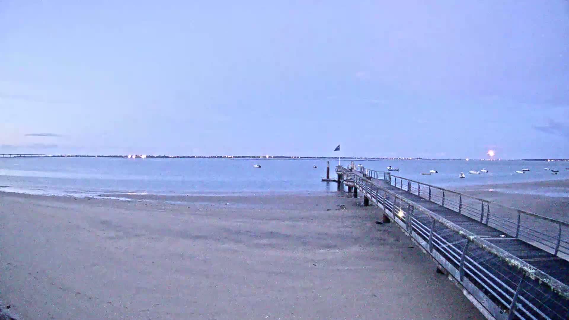 A long pier extends from a sandy beach into a calm body of water under a pale, twilight sky, with several boats visible in the distance.