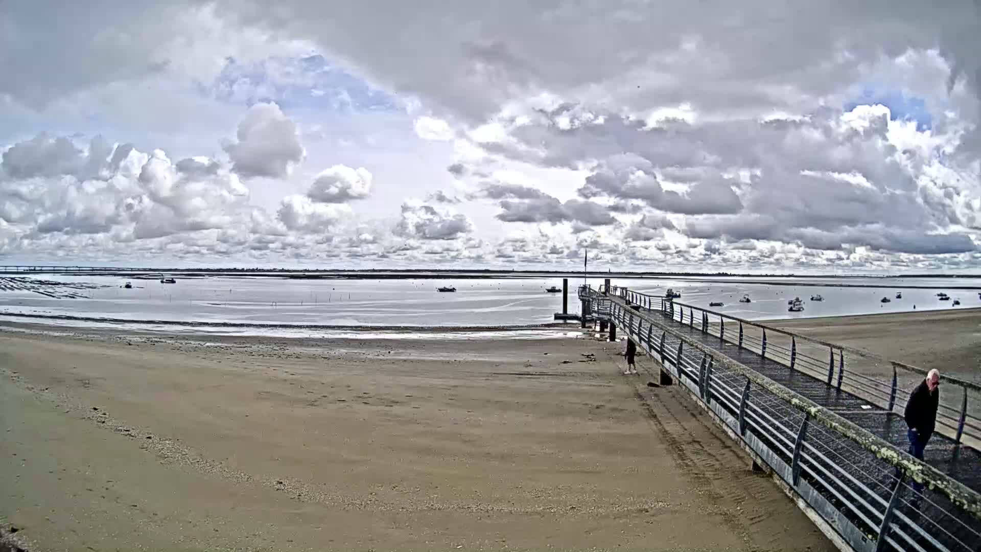 A sandy beach stretches toward a metal pier extending into calm, shallow water under a partly cloudy sky, with several small boats visible in the distance.