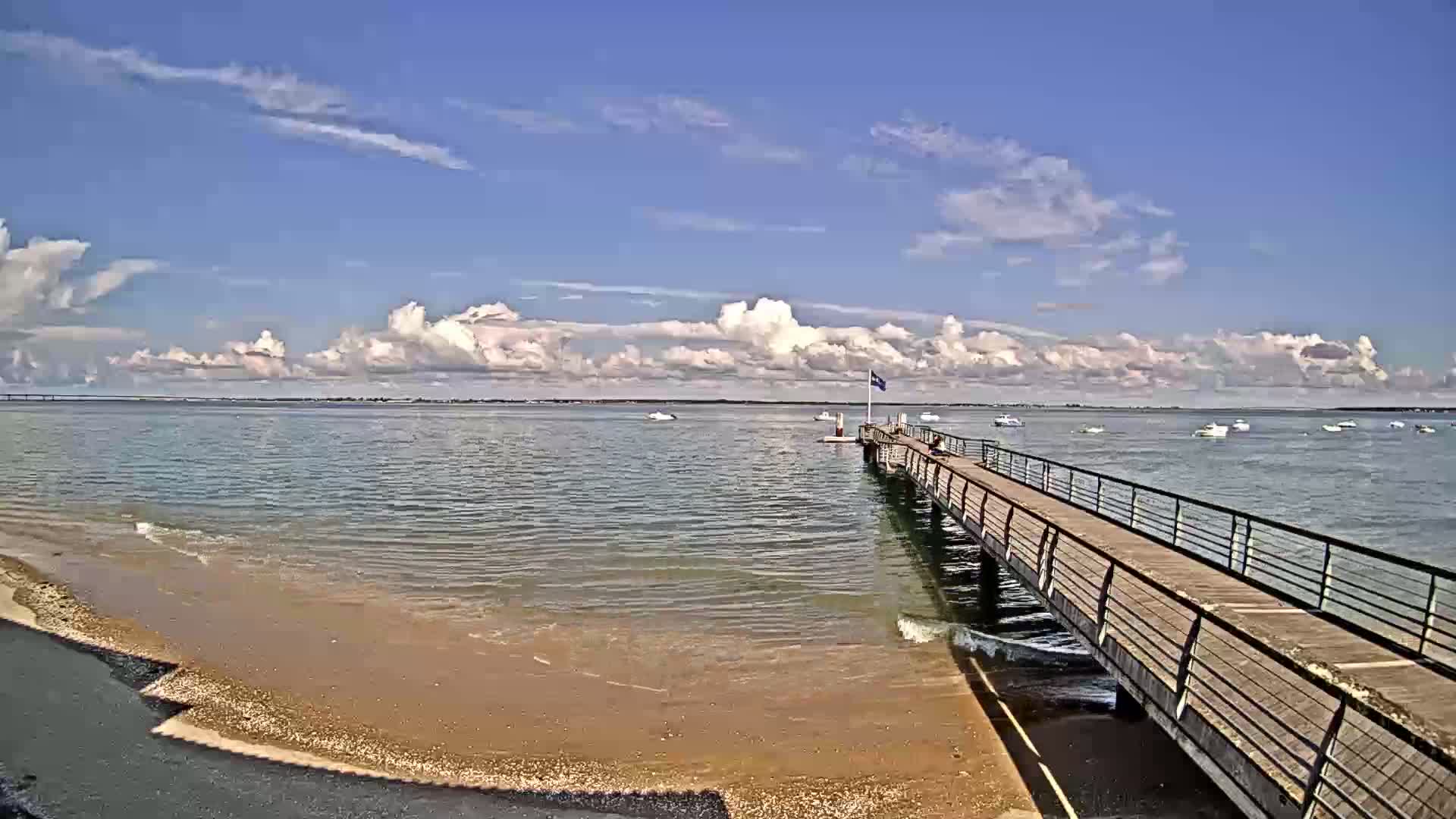 A long pier extends into calm, shallow water under a partly cloudy sky, with several small boats visible in the distance.