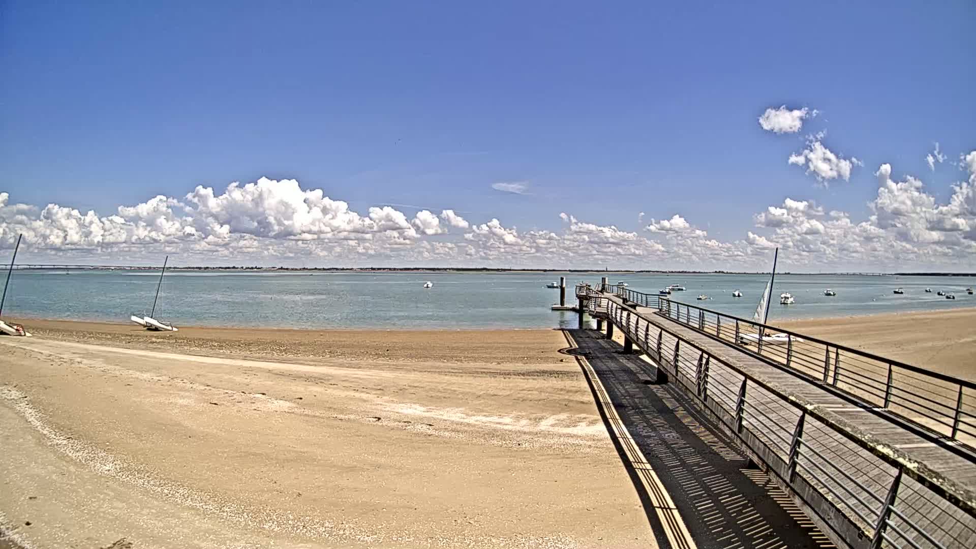 A sandy beach with several small sailboats in the water, a long wooden pier extending from the shore under a partly cloudy blue sky.