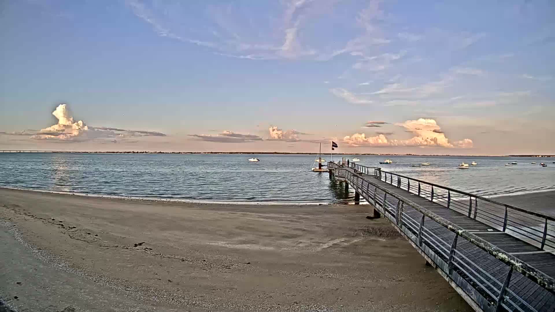 A long pier extends from a sandy beach into calm water under a partly cloudy sky, with several small boats visible in the distance.