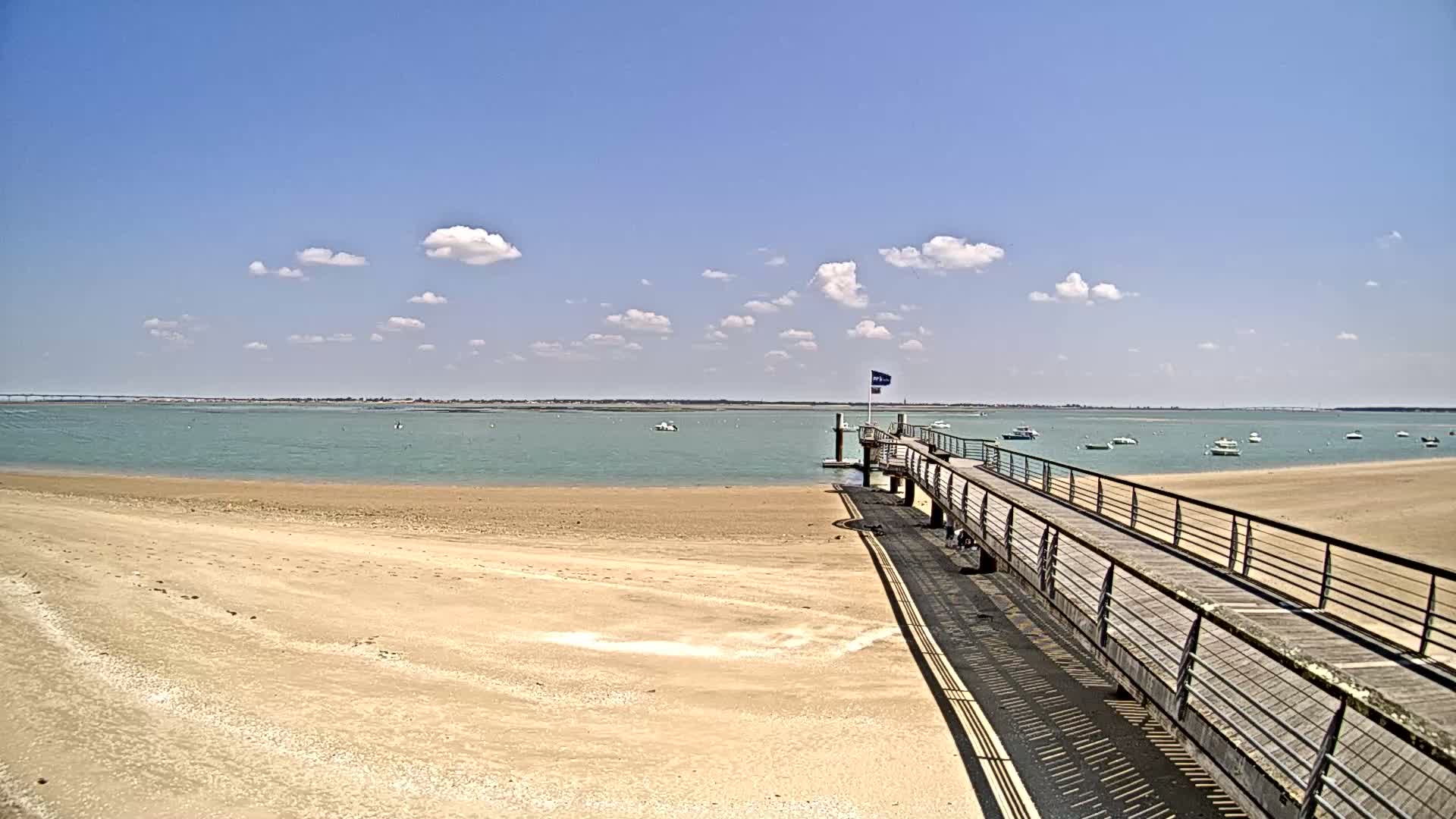 A sandy beach leads to a long wooden pier extending into a calm, light-green body of water under a mostly sunny sky with scattered clouds.