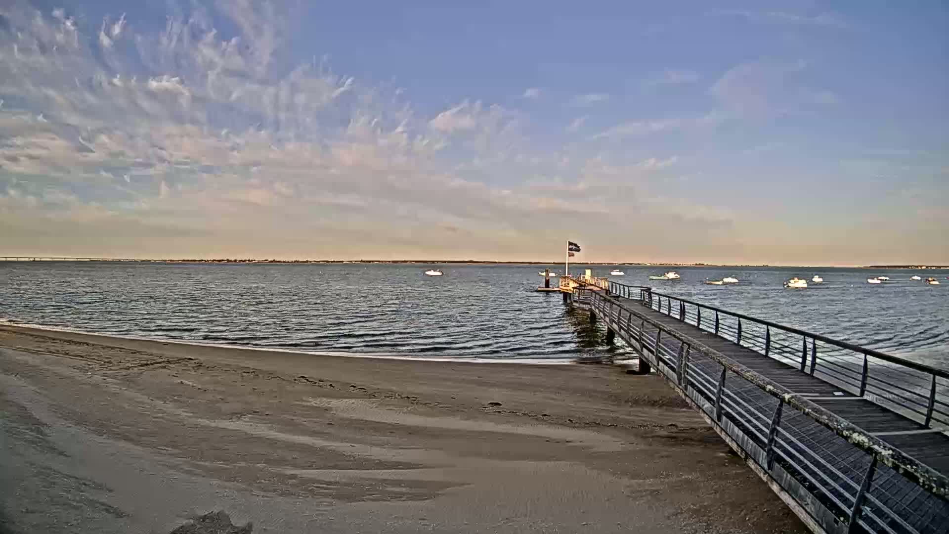 A sandy beach slopes down to a calm body of water where a long pier extends from the shore, under a mostly clear sky with wispy clouds.