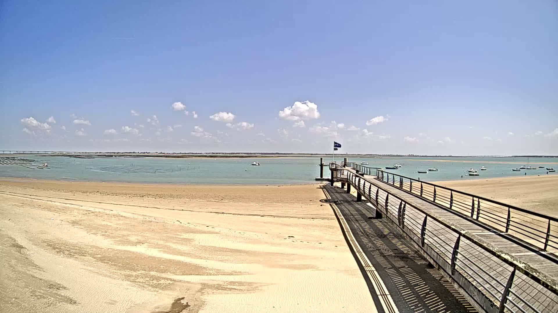 A long wooden pier extends from a sandy beach into a calm, light-blue body of water under a sunny sky with scattered clouds.