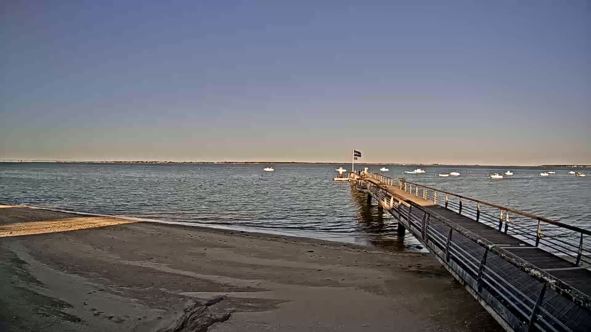 A long pier extends from a sandy beach into calm water, with several small boats visible in the distance under a clear sky.