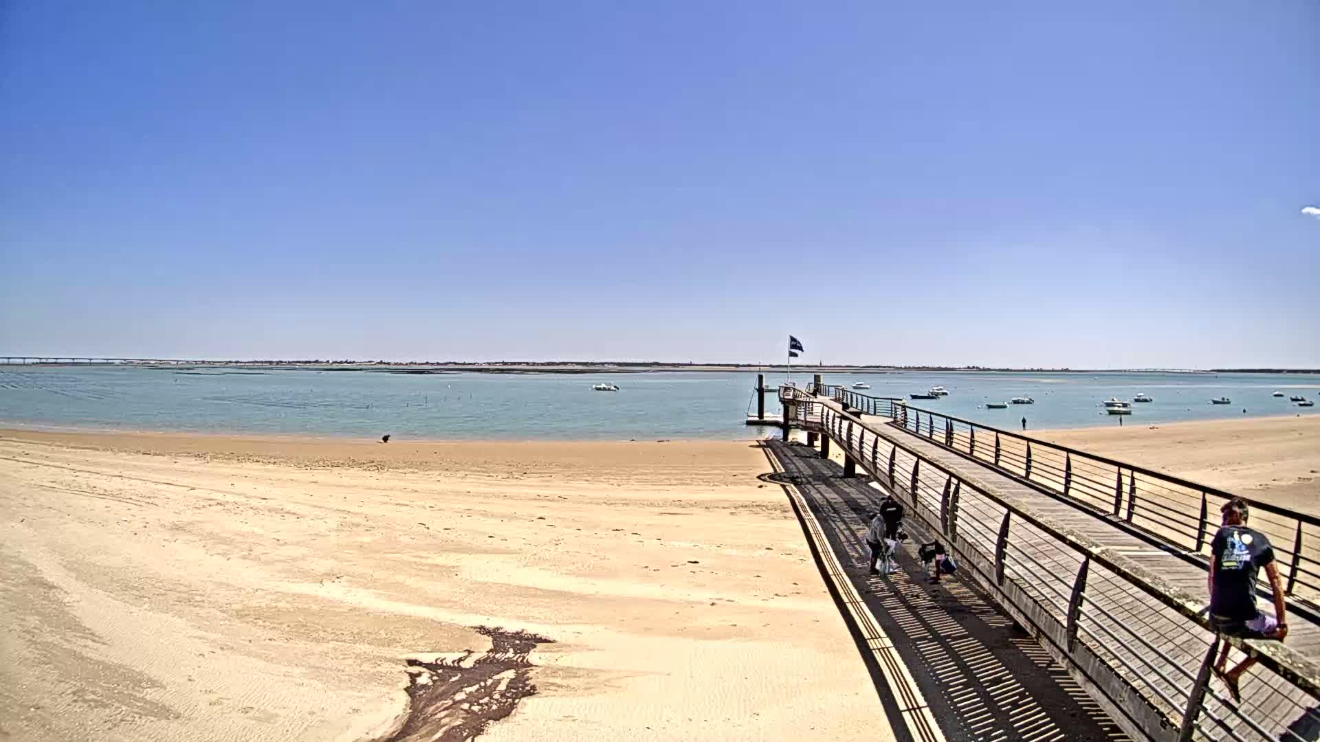 A long wooden pier extends from a sandy beach into calm, shallow water dotted with small boats under a clear blue sky.