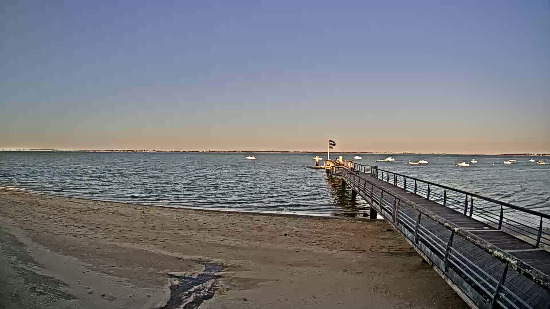 A long pier extends from a sandy beach into calm water where several small boats are anchored, under a clear sky.