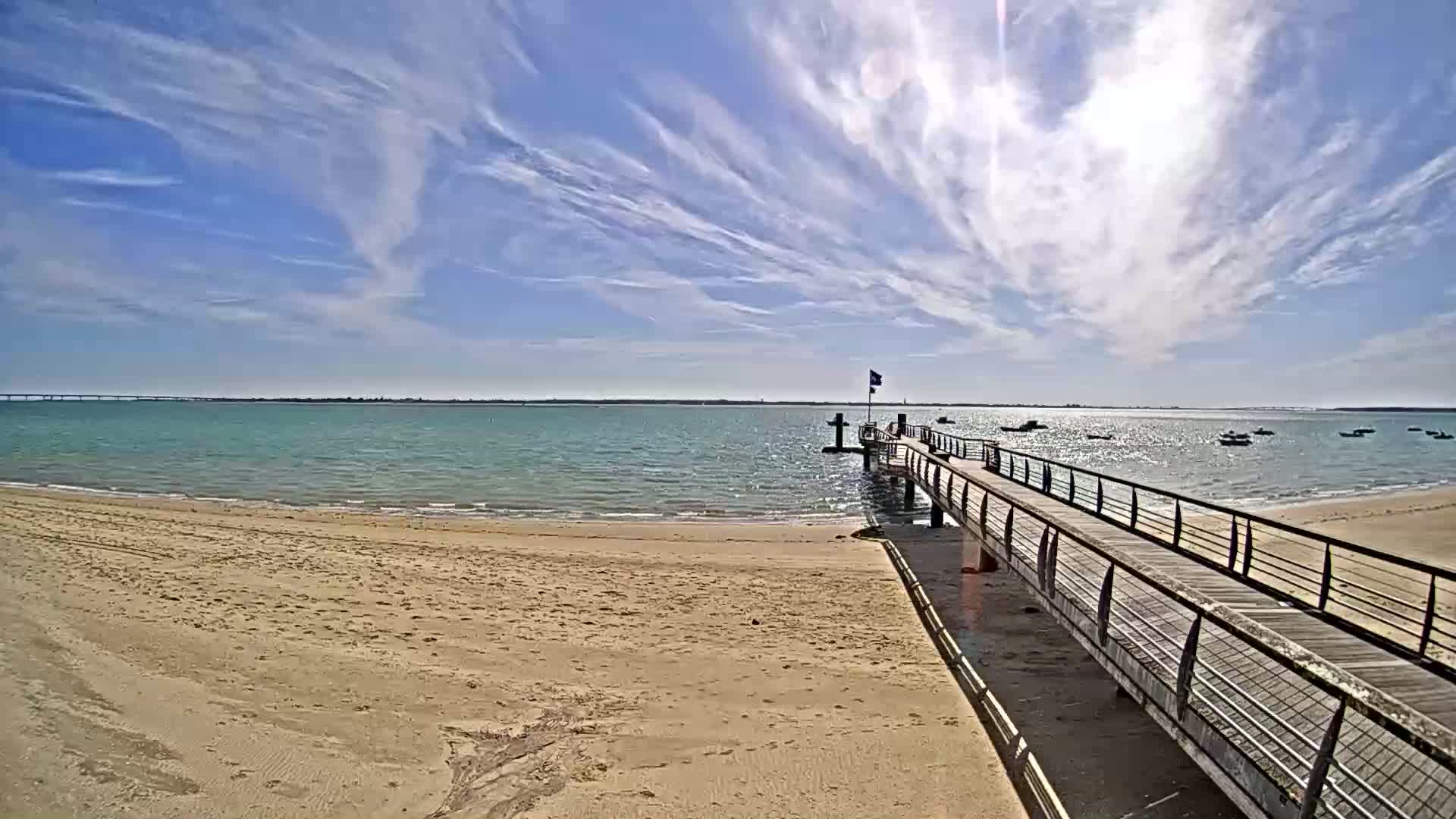 A sandy beach leads to a wooden pier extending into a calm, light-blue body of water under a mostly sunny sky with thin, wispy clouds.