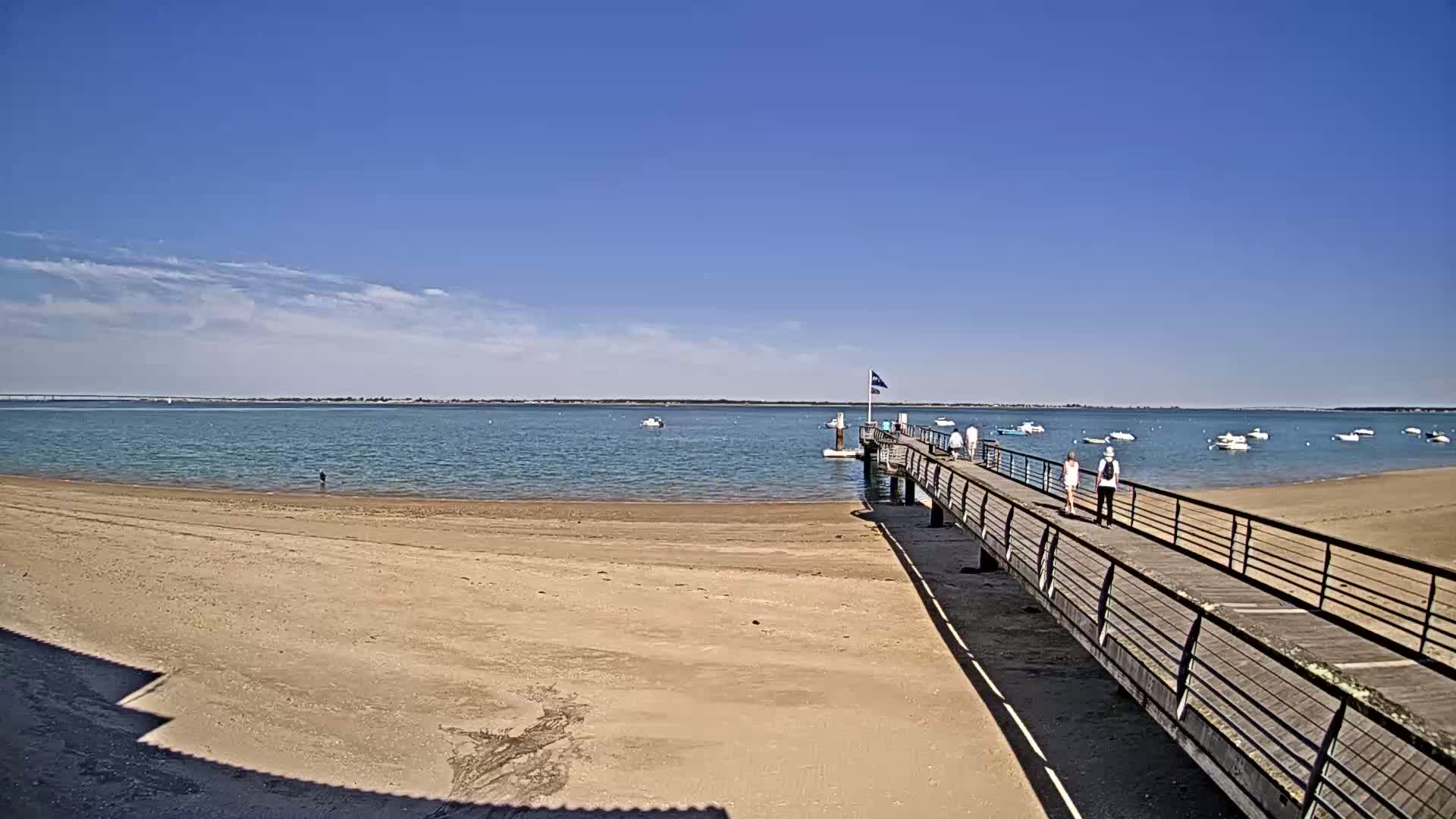 A sandy beach stretches out to a calm, blue body of water where several small boats are anchored; a long pier extends from the beach with a few people walking along it under a clear, sunny sky.