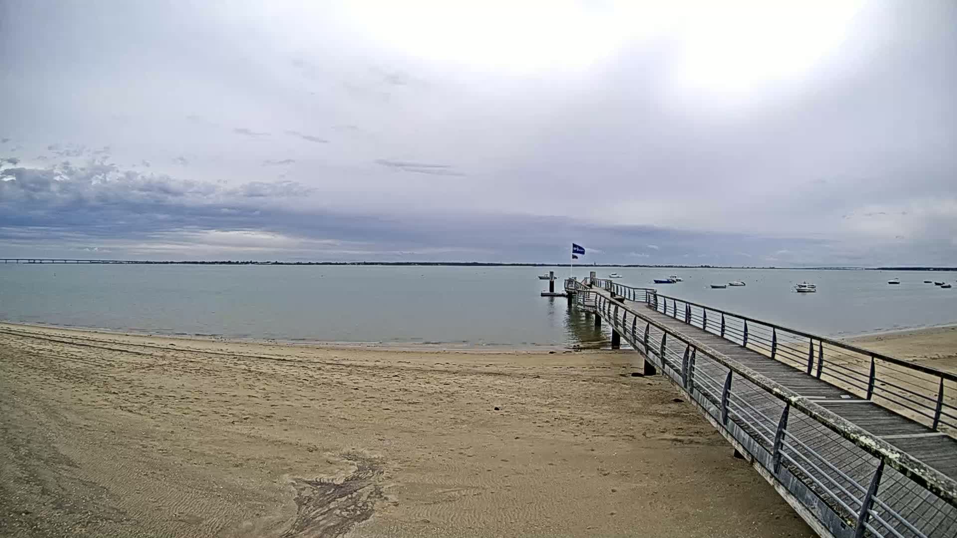 A sandy beach stretches towards a calm body of water where a long pier extends from the shore under an overcast sky, with several small boats visible in the distance.