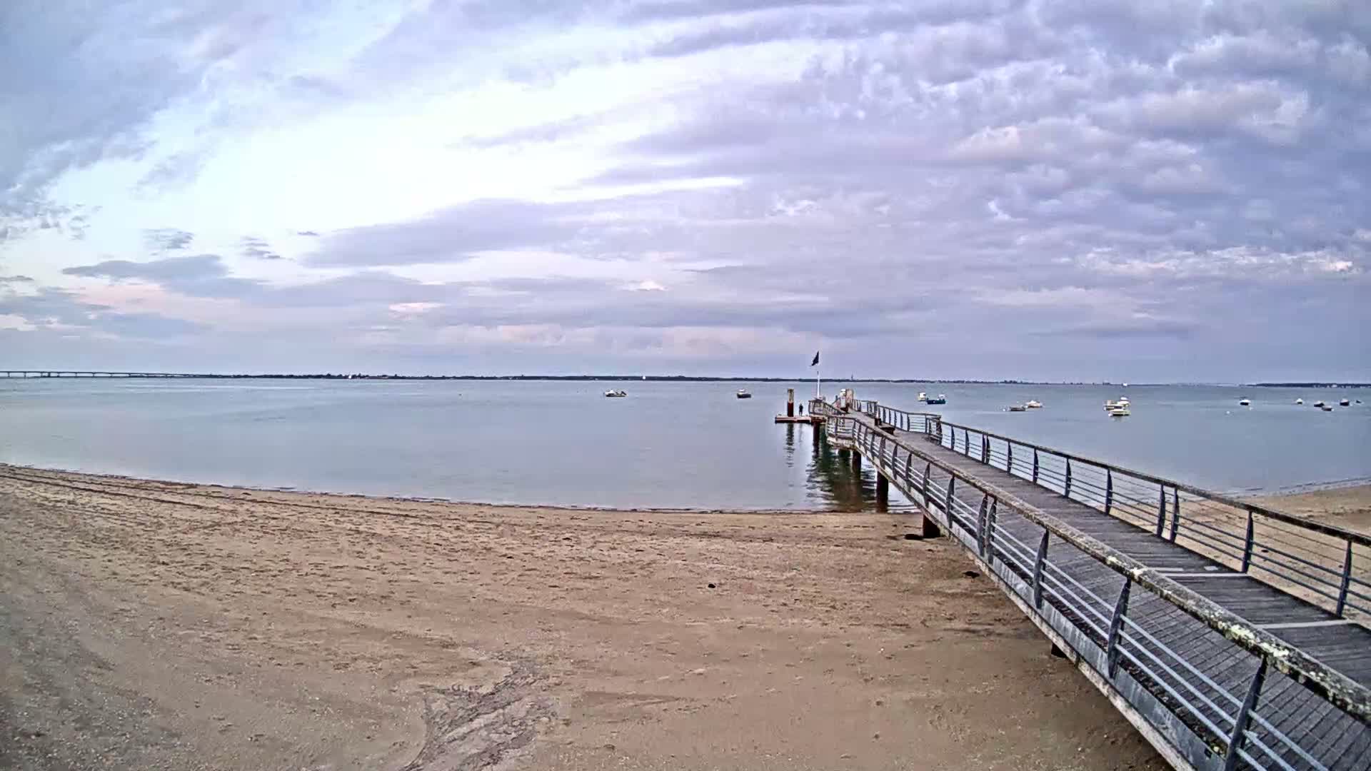A long pier extends from a sandy beach into calm water under a cloudy sky, with several small boats visible in the distance.