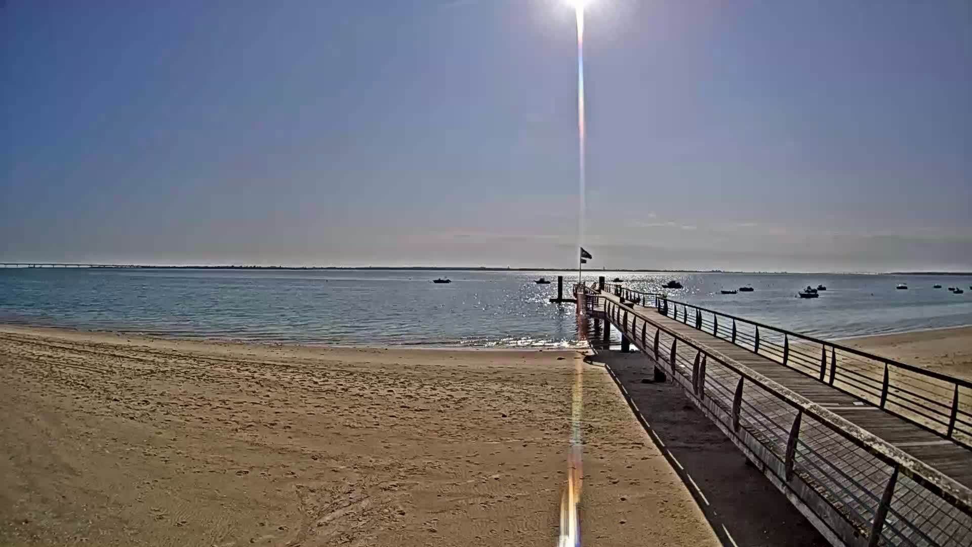 A long wooden pier extends from a sandy beach into calm, sunlit water dotted with small boats under a clear blue sky.