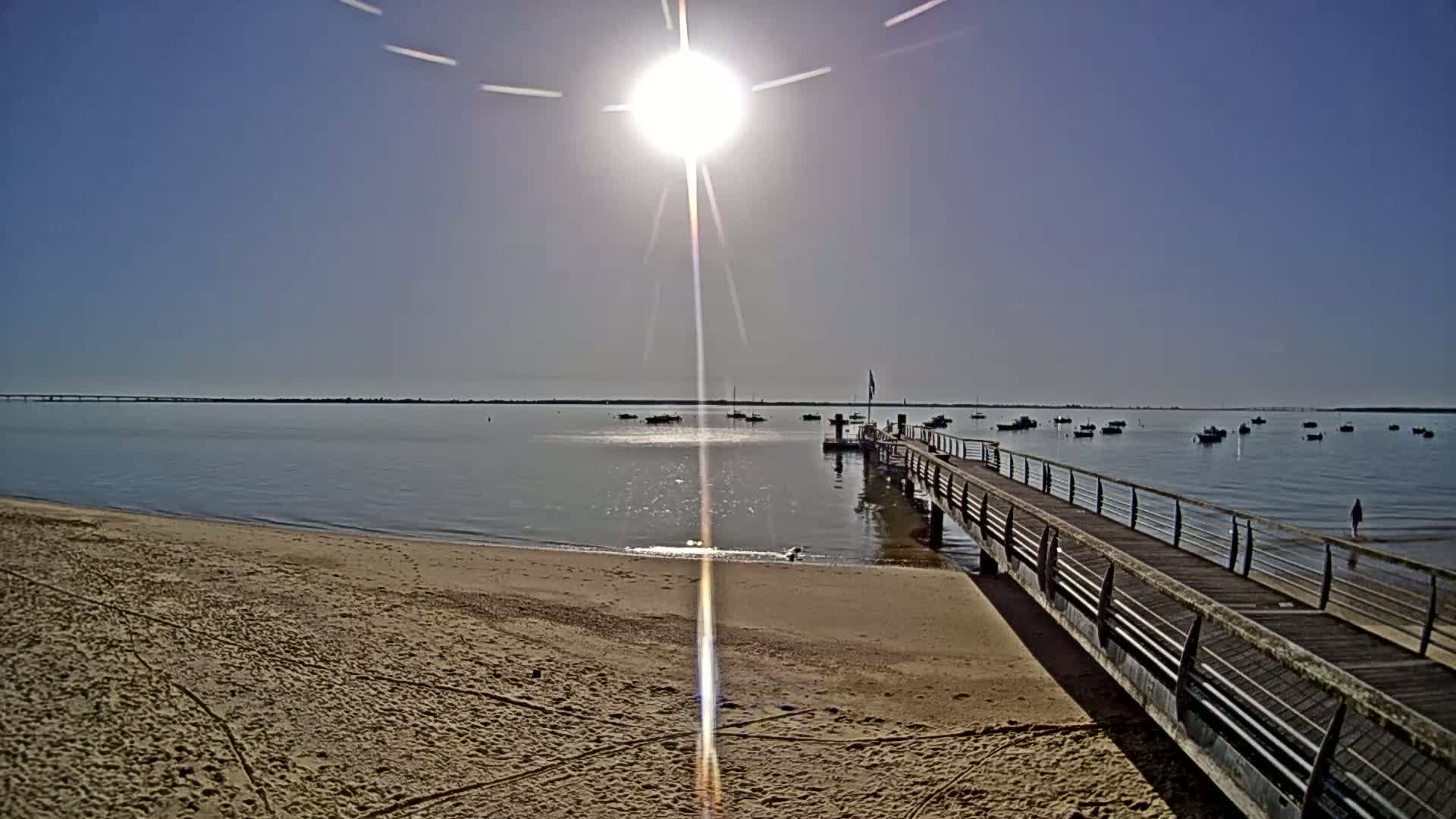 A sandy beach with a long pier extends into a calm body of water under a bright, sunny sky, where several small boats are anchored.