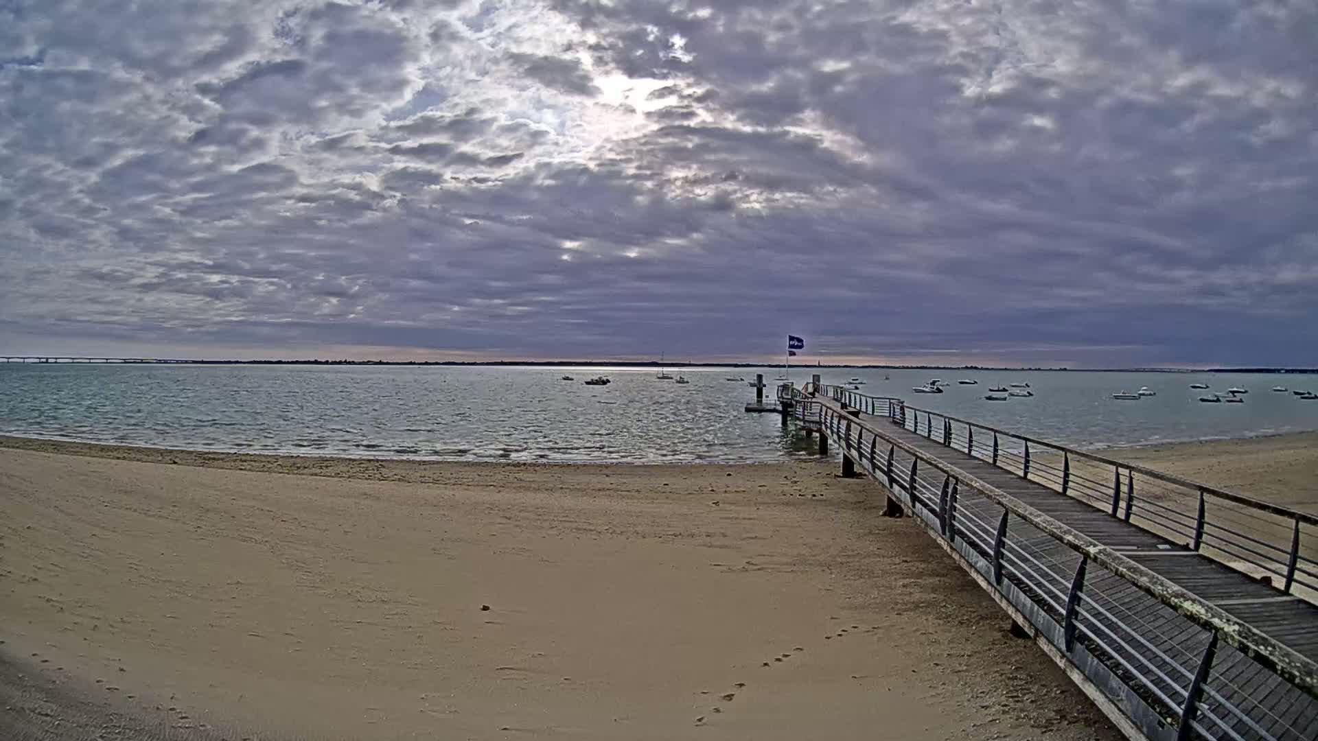 A sandy beach leads to a wooden pier extending into calm water under a cloudy sky, with several small boats visible in the distance.