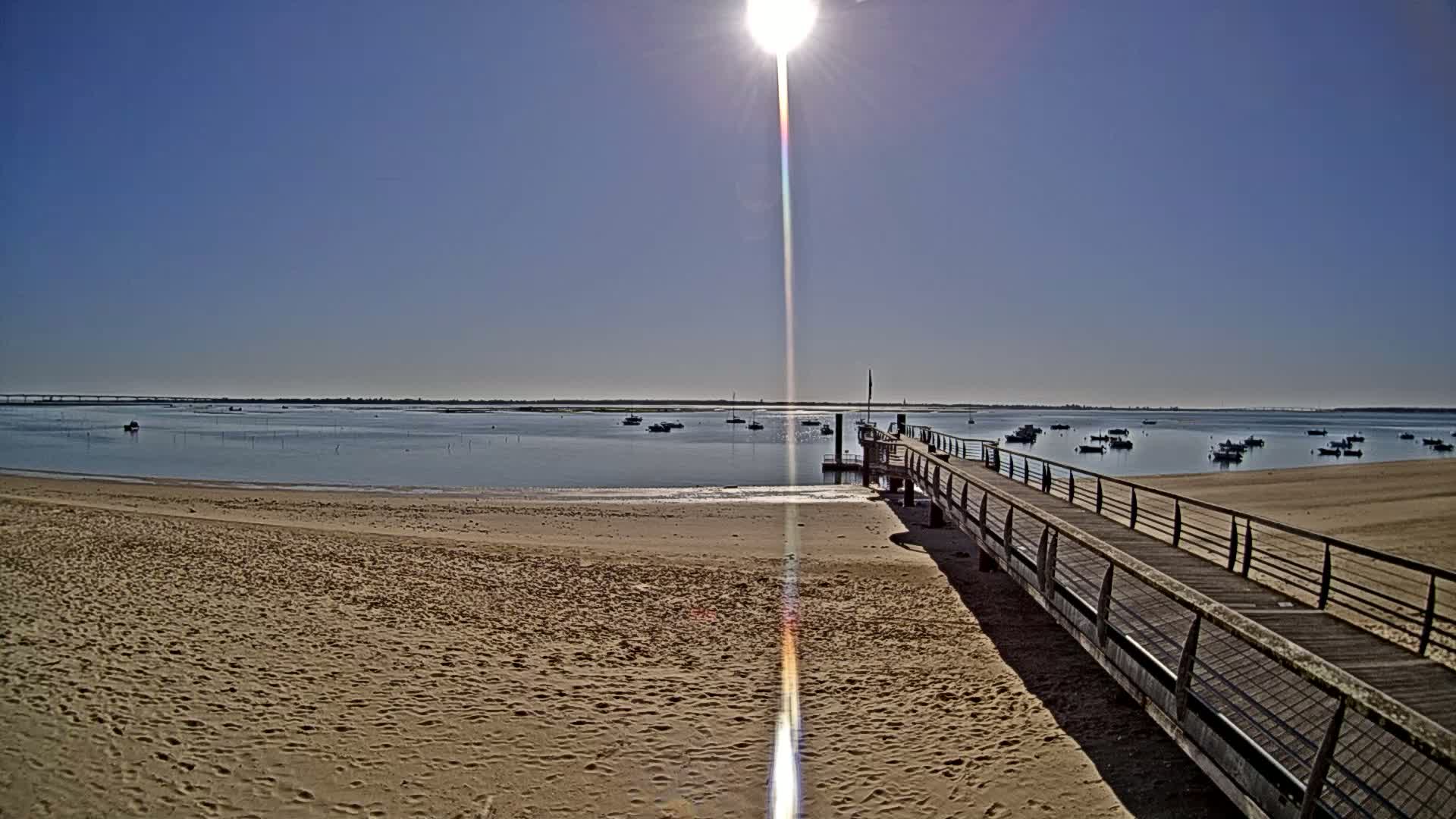 A sandy beach stretches towards a calm body of water with several boats, under a bright, sunny sky, and a long wooden pier extends from the shore.