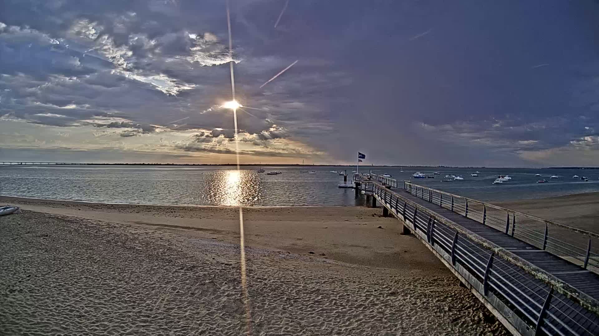 A sandy beach with a long pier extends into a calm body of water under a partly cloudy sunset sky.