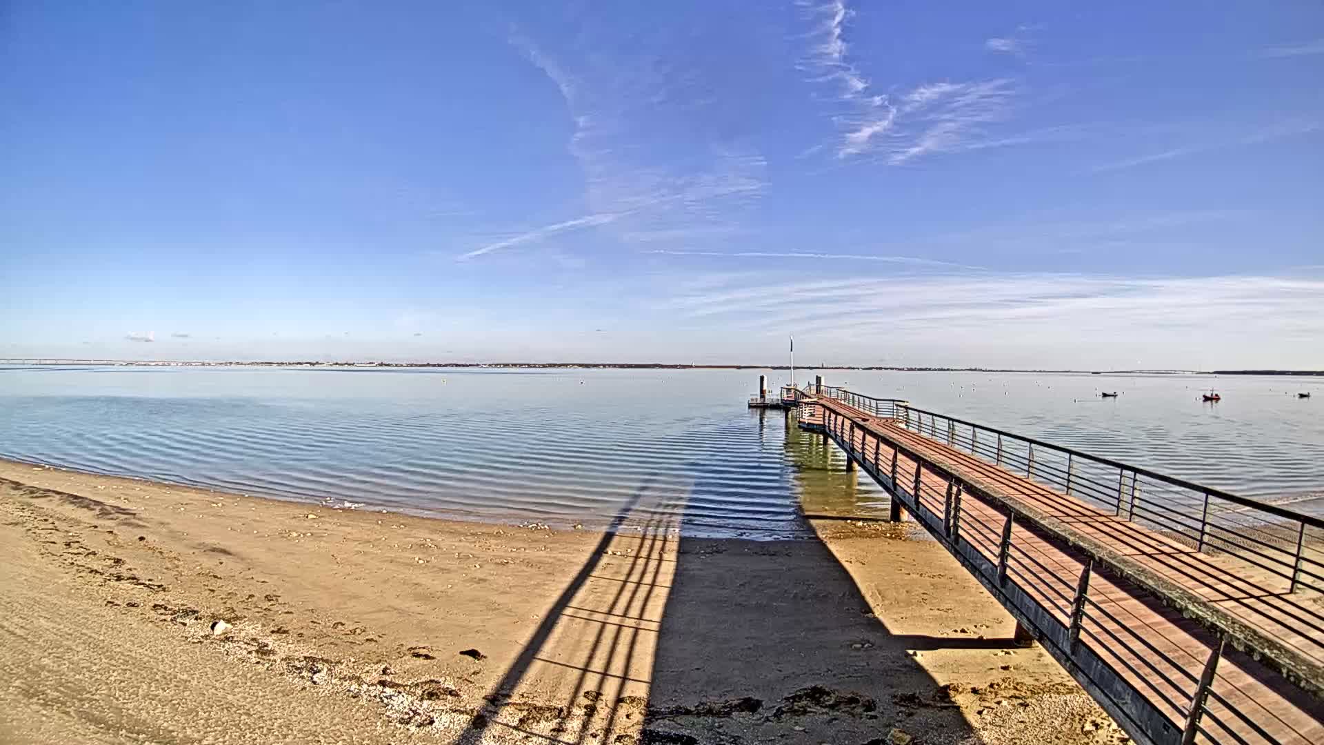 A long wooden pier stretches over a sandy beach and calm bay waters towards a flagpole, all under a uniformly cloudy sky with a distant bridge and shoreline visible on the horizon.