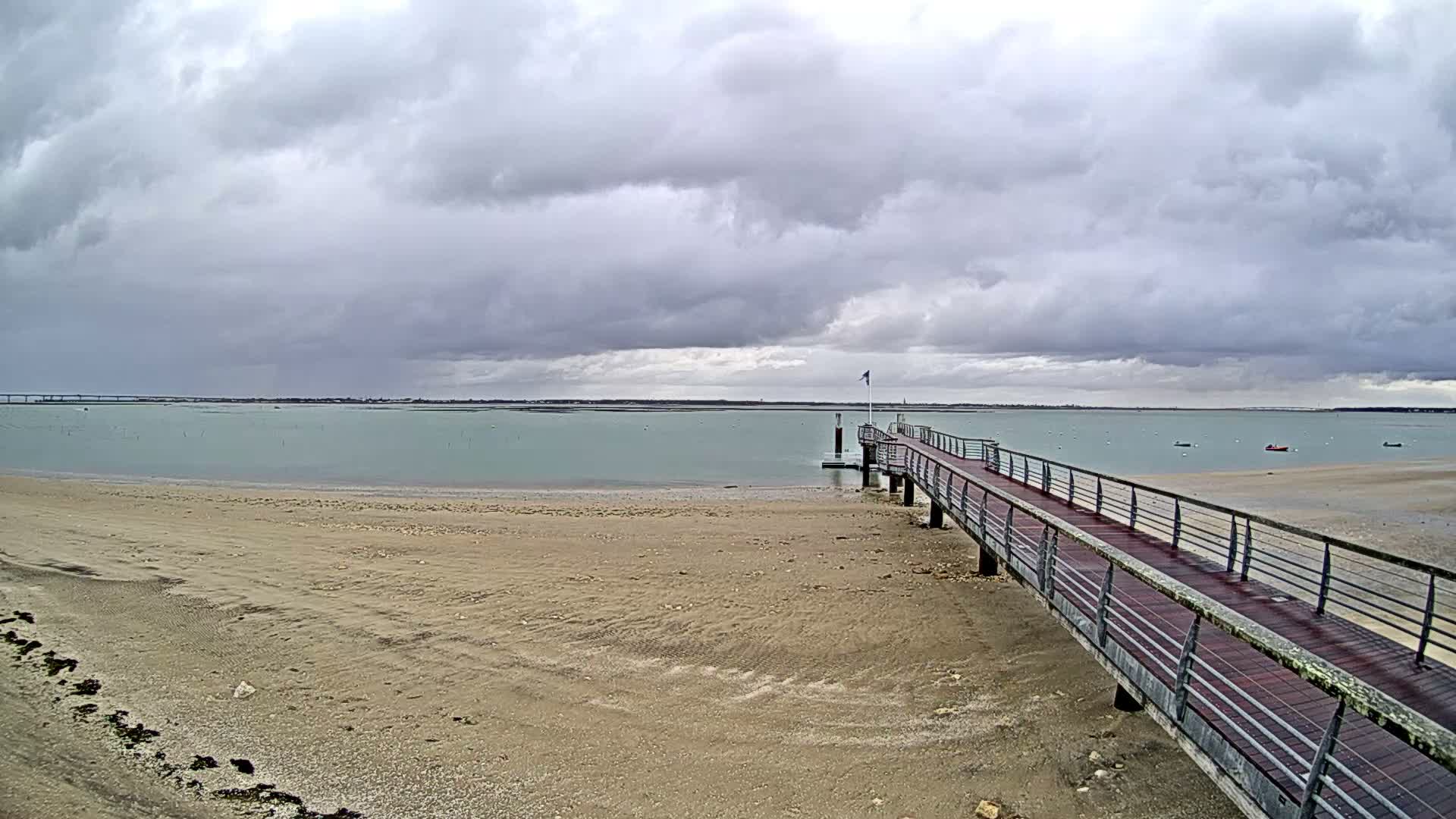A long wooden pier with metal railings stretches from a wide sandy beach into calm, light blue-green water, all beneath a heavily overcast sky.