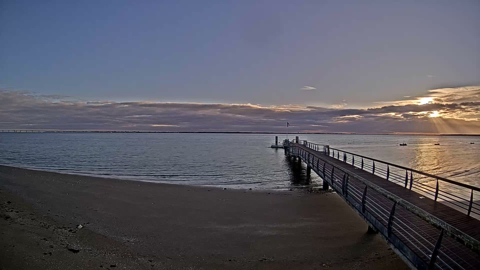 A long wooden pier extends into calm water from a sandy beach under a partly cloudy sky with sunbeams breaking through on the right horizon, revealing a distant bridge to the left.