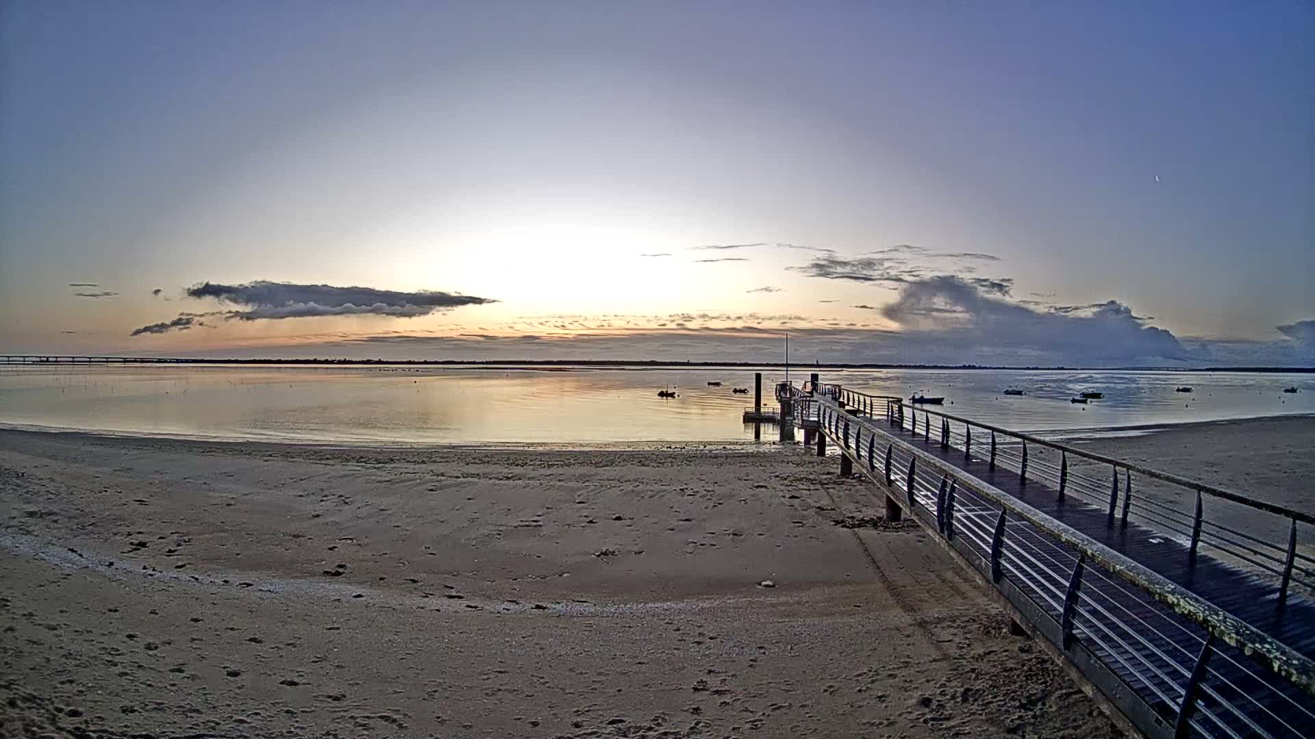 A long wooden pier extends into calm water from a sandy beach under a partly cloudy sky with sunbeams breaking through on the right horizon, revealing a distant bridge to the left.