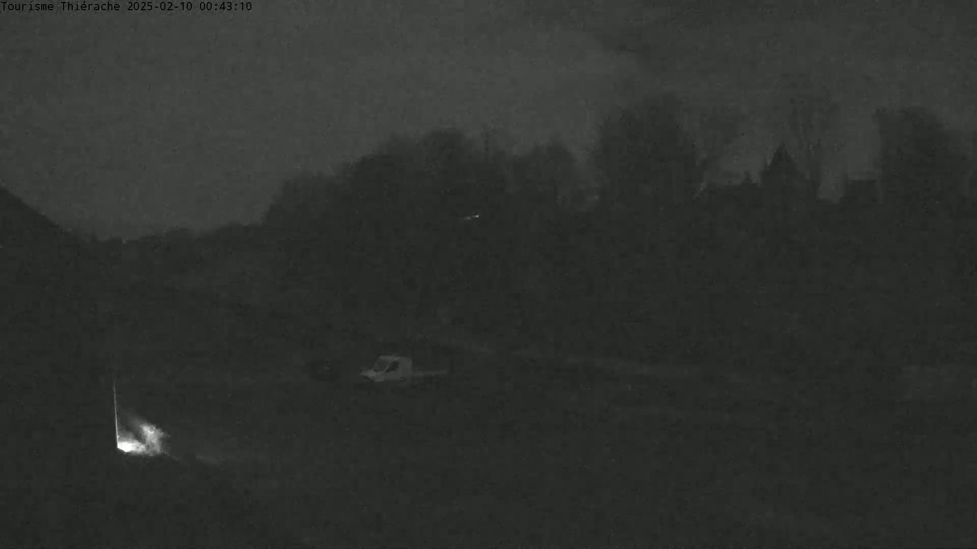 A nighttime, low-light image shows a dark landscape with a few indistinct buildings and vehicles under a cloudy sky.