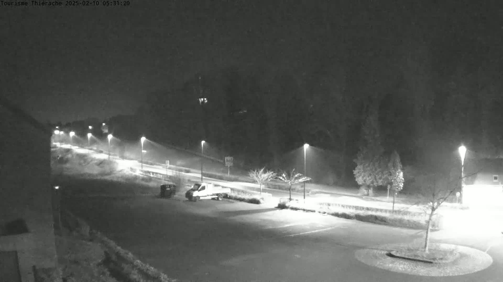 A nighttime, snow-covered parking lot and road are illuminated by streetlights, with a few vehicles visible.