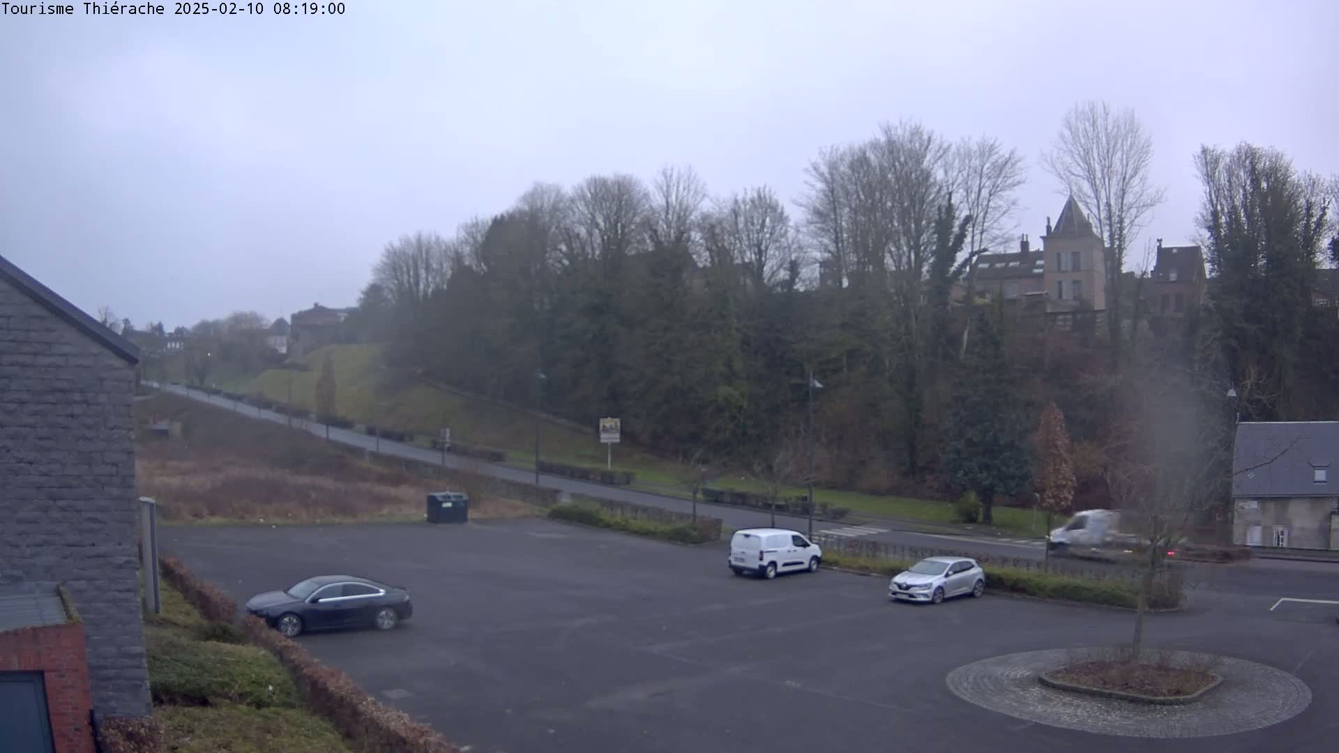 A mostly empty parking lot with a few cars and a van, overlooks a road that leads to a hill topped with buildings and trees under an overcast sky.