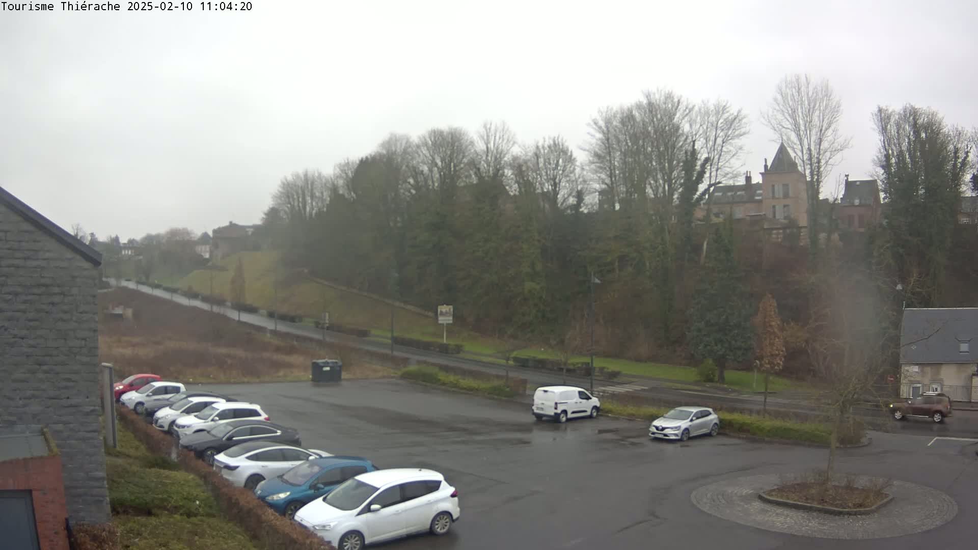 A parking lot with several cars is seen next to a road leading to a hill covered in bare trees and buildings, all under an overcast sky.