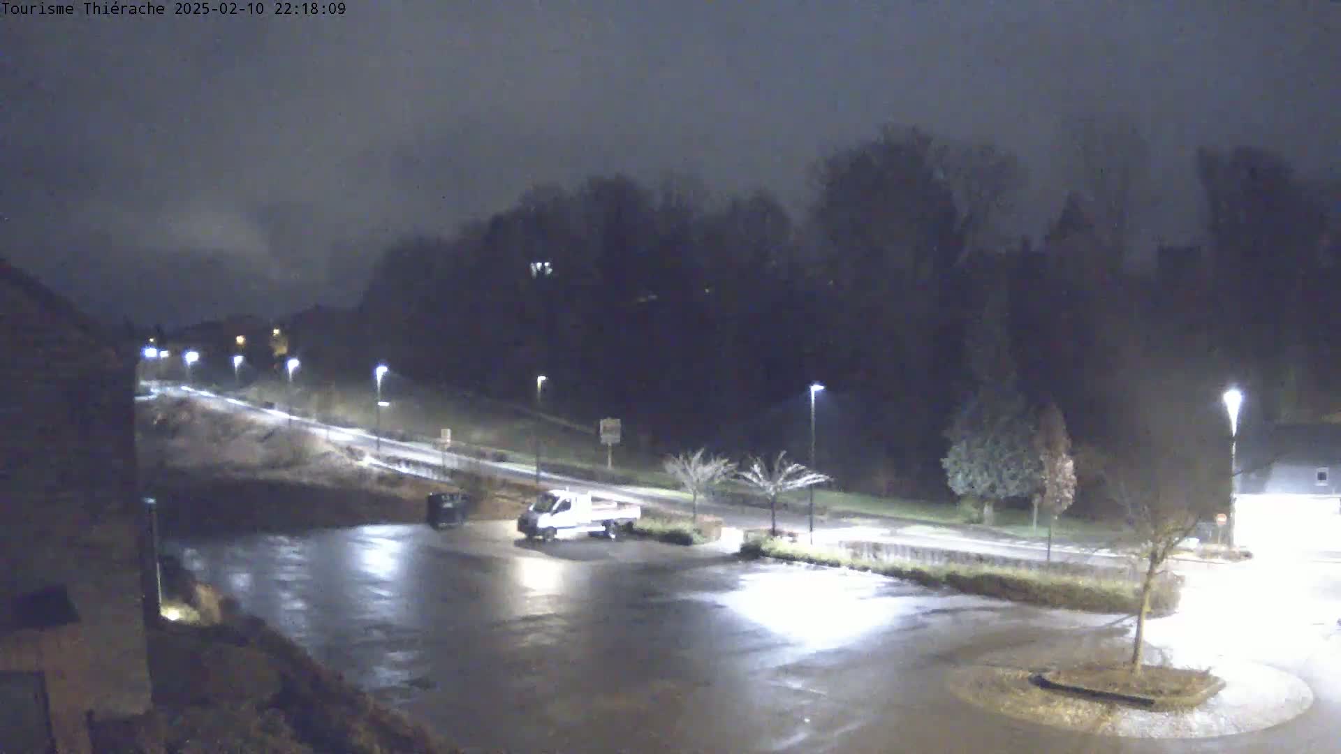 A mostly empty, wet parking lot at night is illuminated by streetlights, with a single van parked nearby and a road leading towards a dark, tree-lined hill in the background.