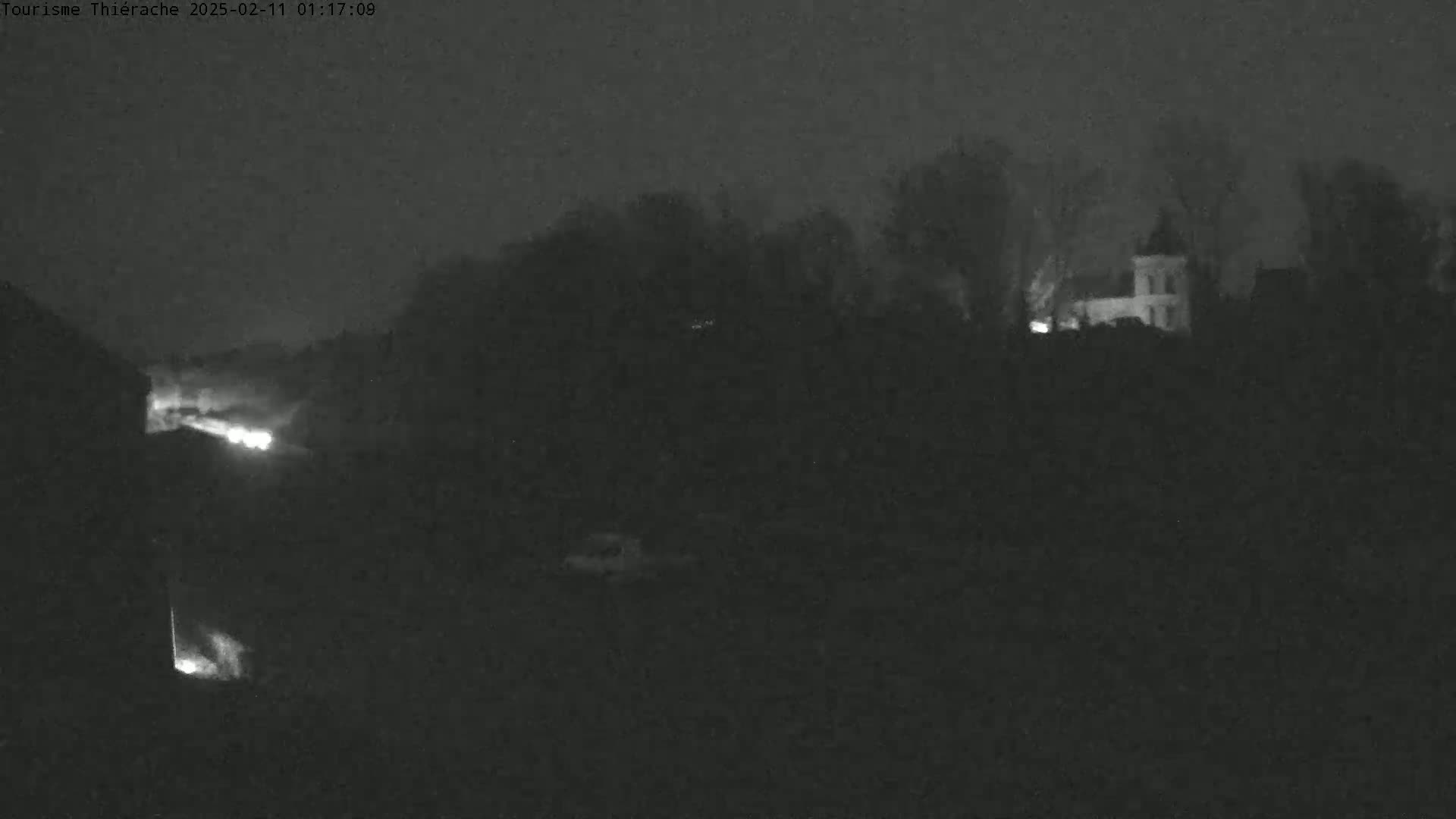 A dimly lit nighttime scene shows a building faintly illuminated among trees, with a bright light source visible in the lower left, under a dark, overcast sky.
