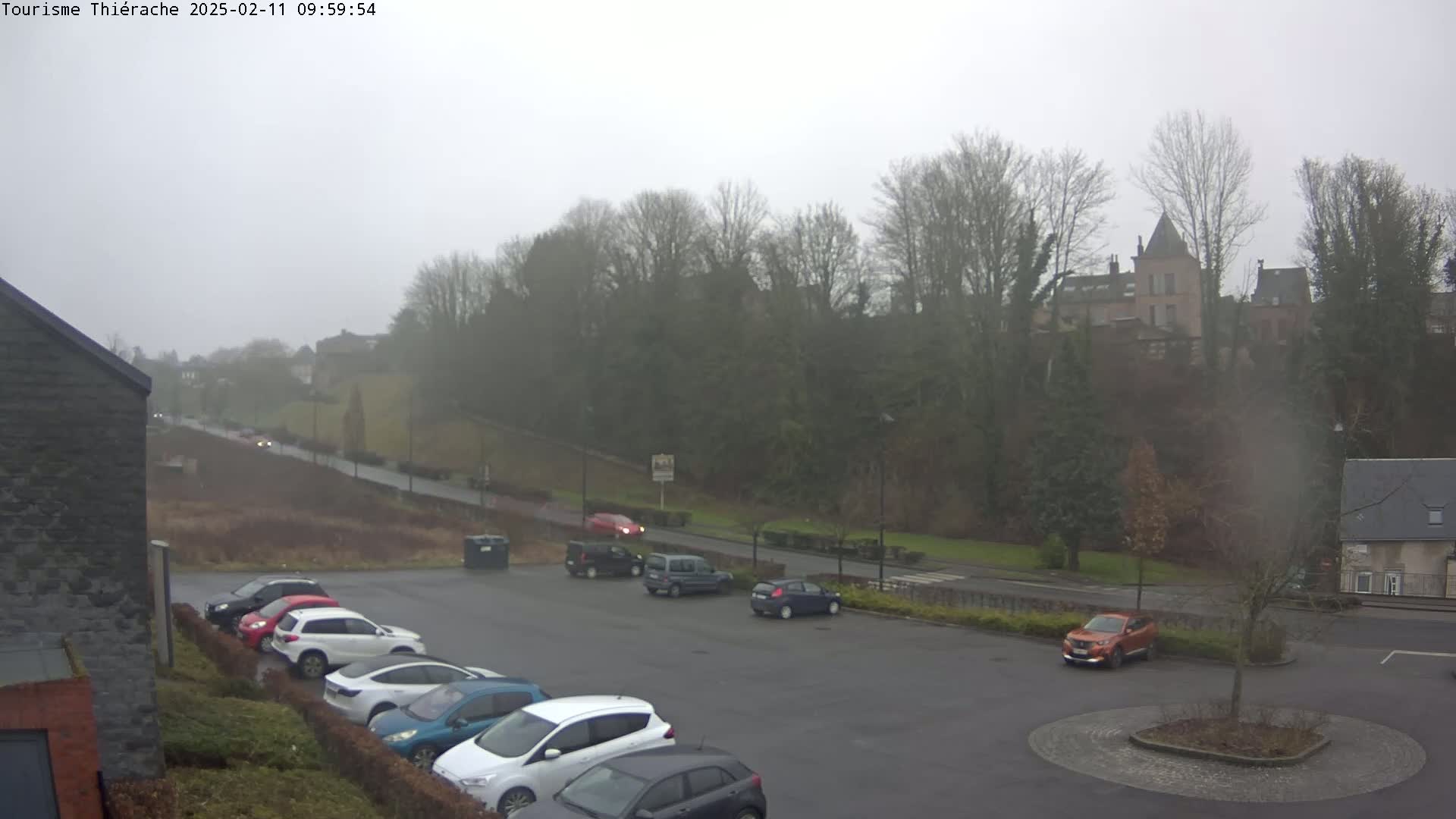 A parking lot with several cars is visible in the foreground, next to a road where additional vehicles are driving; beyond the road, a hill covered with trees and a building is seen under an overcast sky.