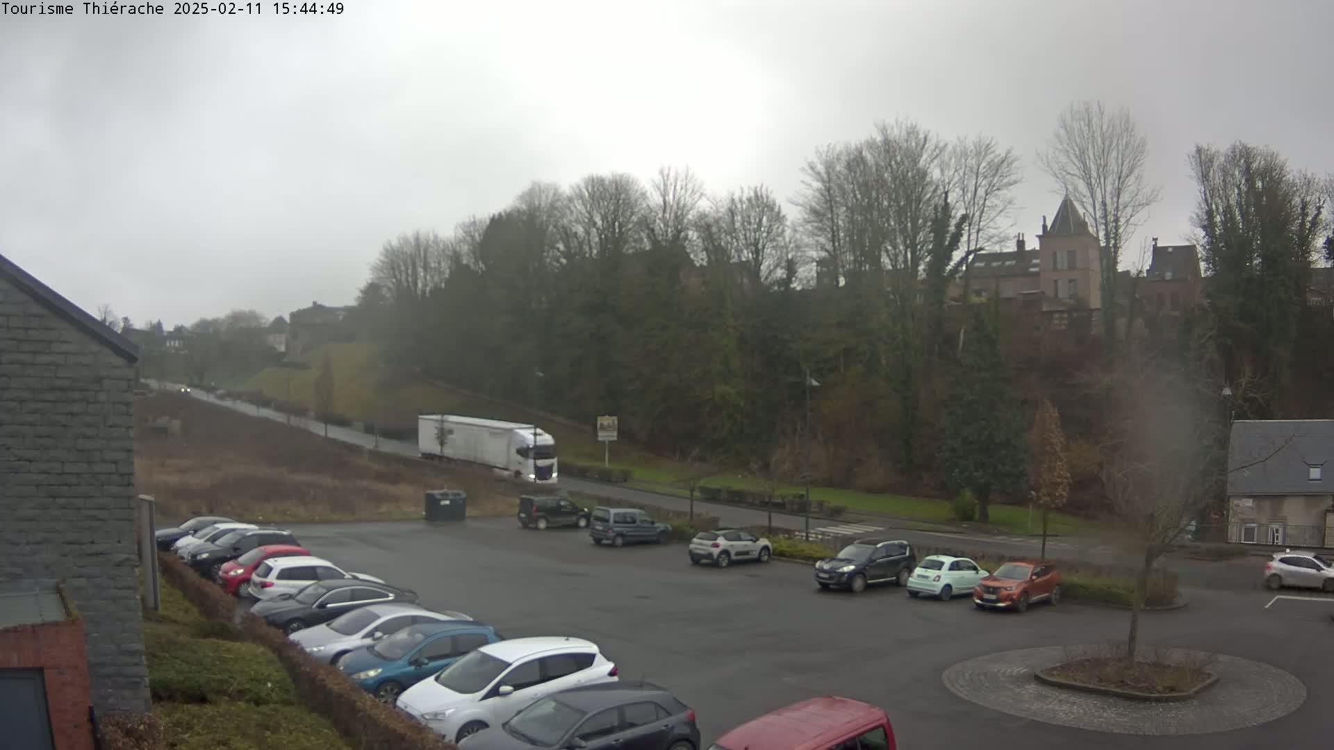 A parking lot with numerous cars is situated below a hill covered in leafless trees and a building, all under an overcast sky.